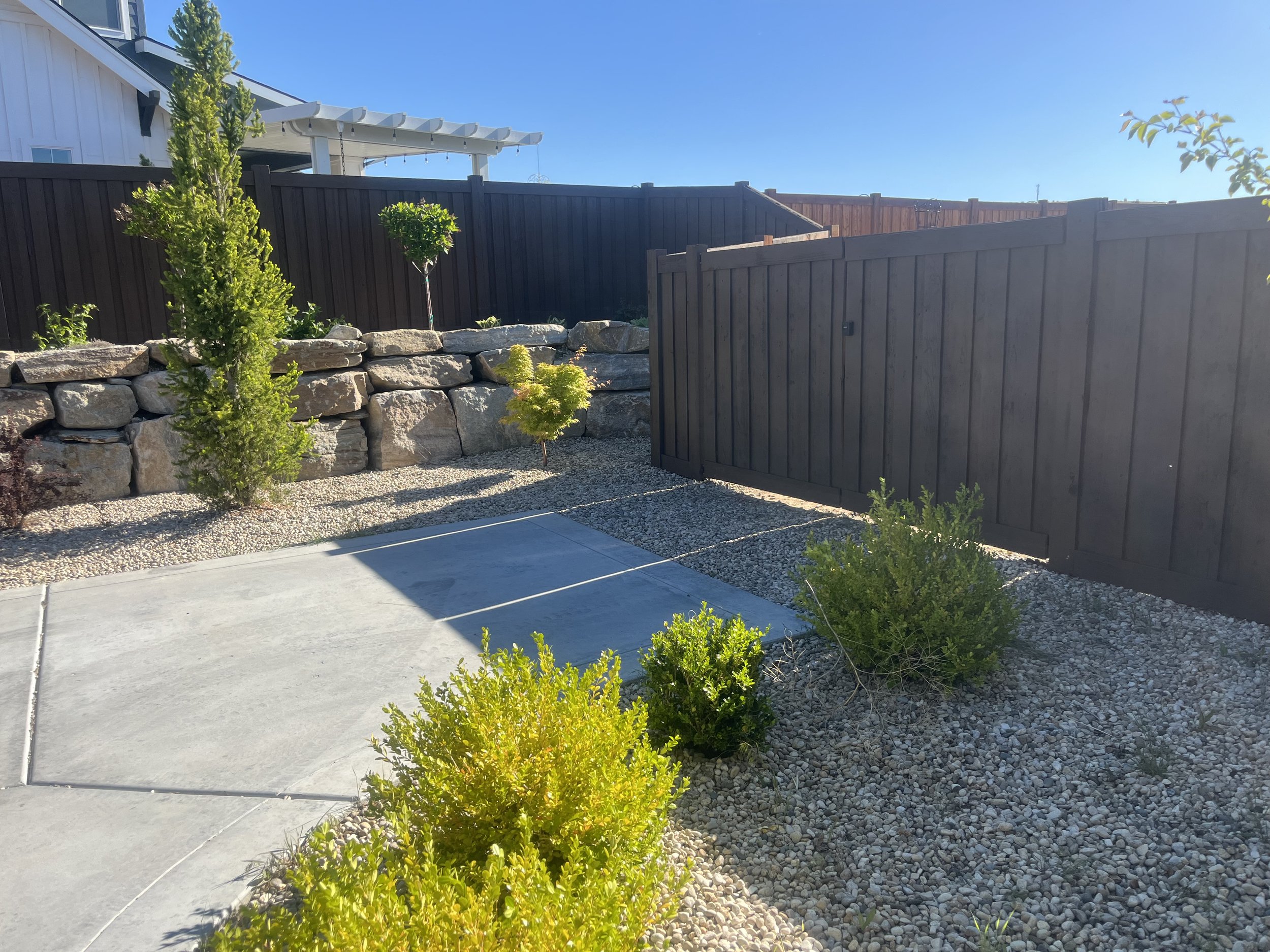 A backyard with a concrete patio, small bushes, a rock retaining wall, and a dark wooden fence under a clear blue sky.