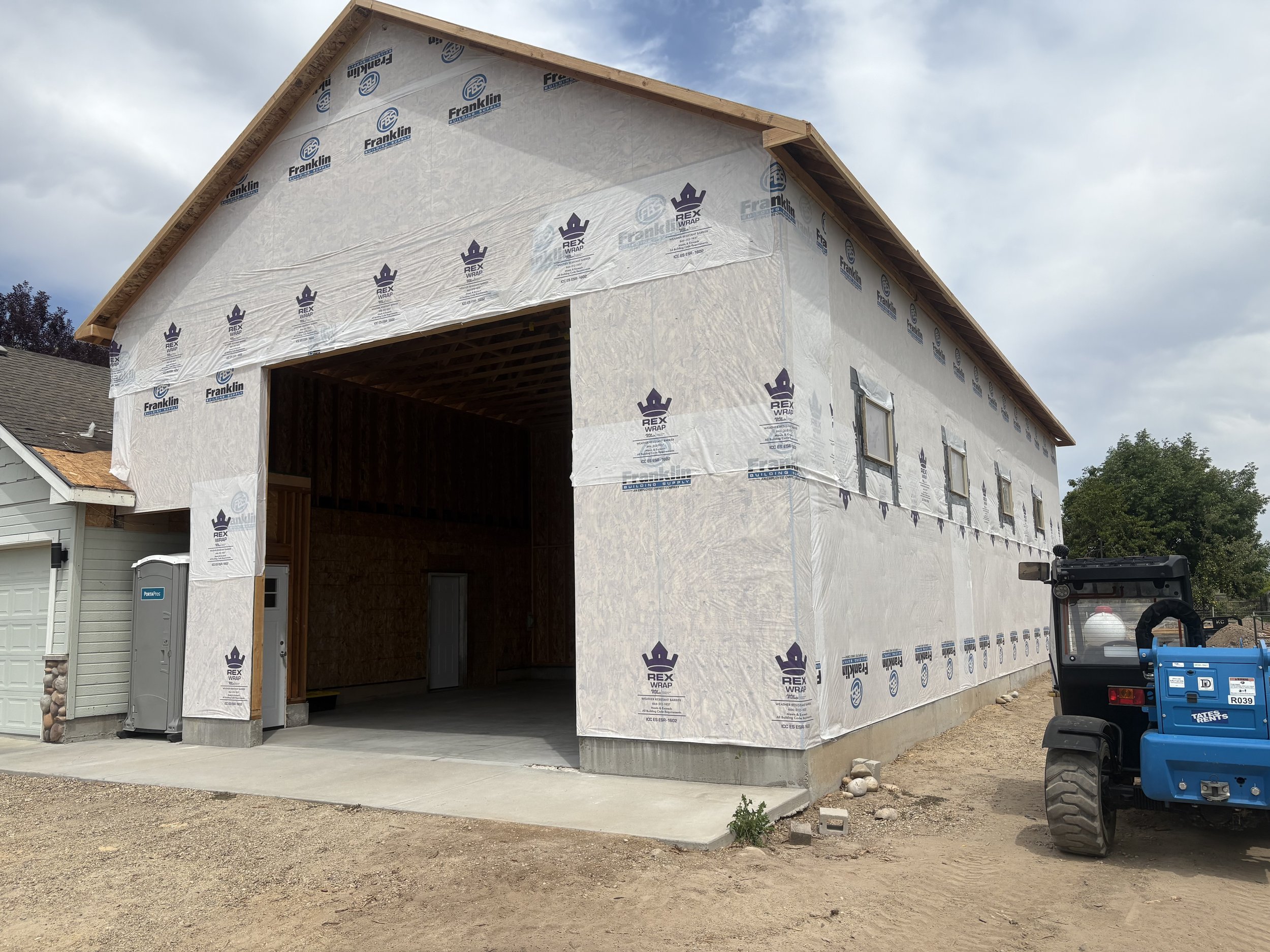 A house under construction with framing and exterior sheathing, covered in protective wrap, and a garage opening with a concrete driveway, on a partly cloudy day.