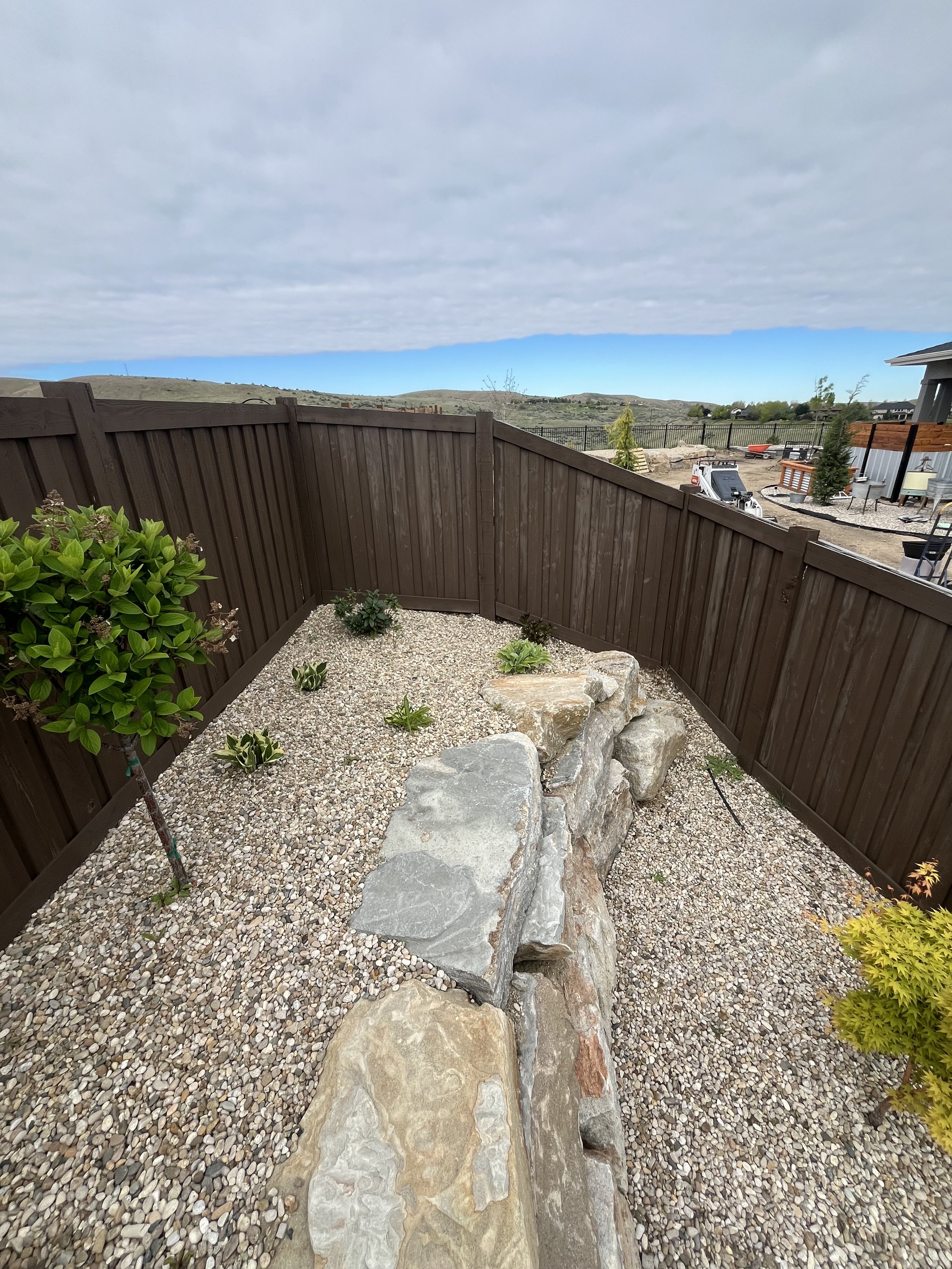 Small backyard with gravel ground, a wooden fence, a few plants, and large rocks, with a cloudy sky and a view of nearby hills and other houses in the background.