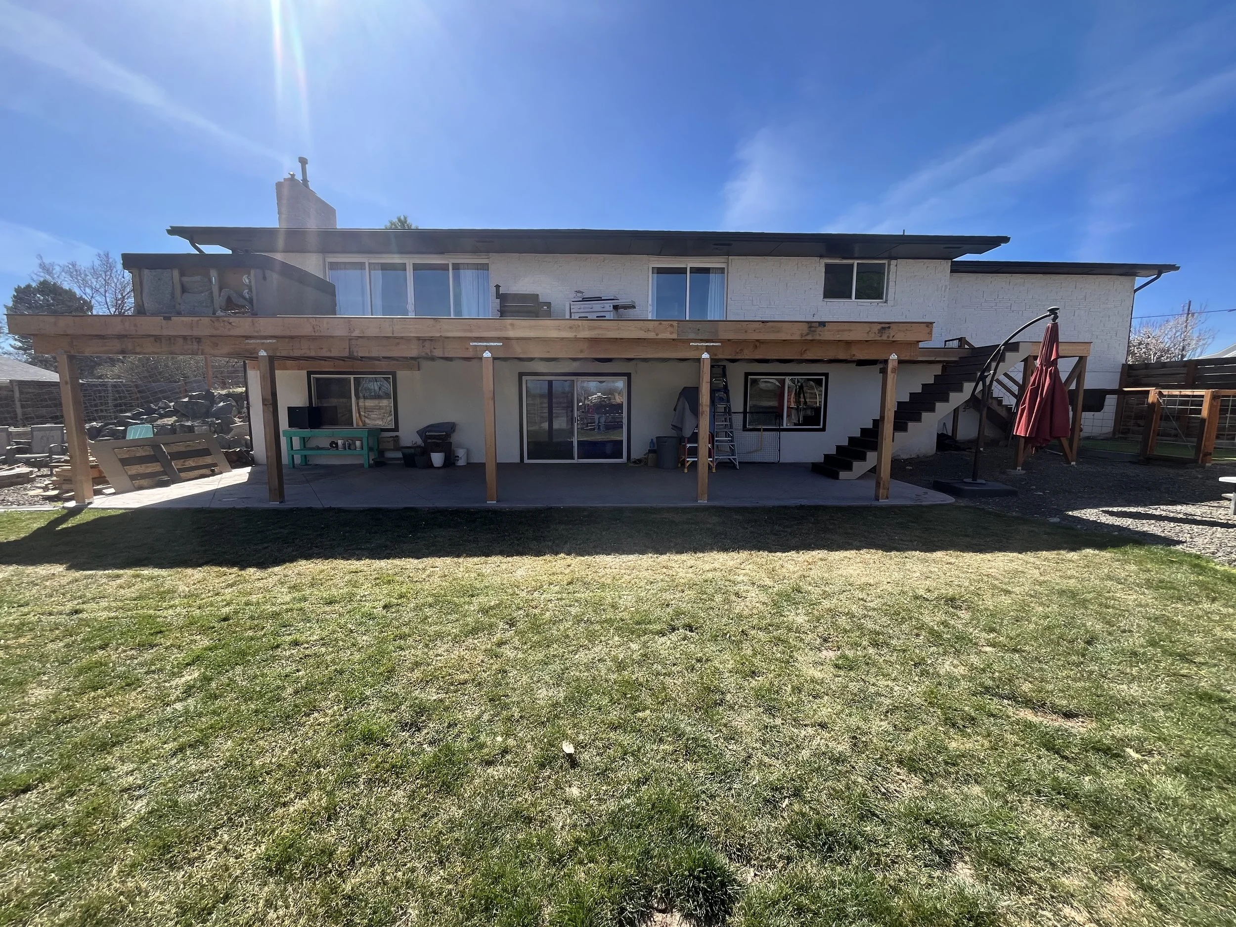 Backyard view of a two-story house with a large unfinished wooden deck on the ground level and a staircase leading to the upper floor. The yard has a grassy lawn, and the photo is taken on a sunny day with a clear blue sky.