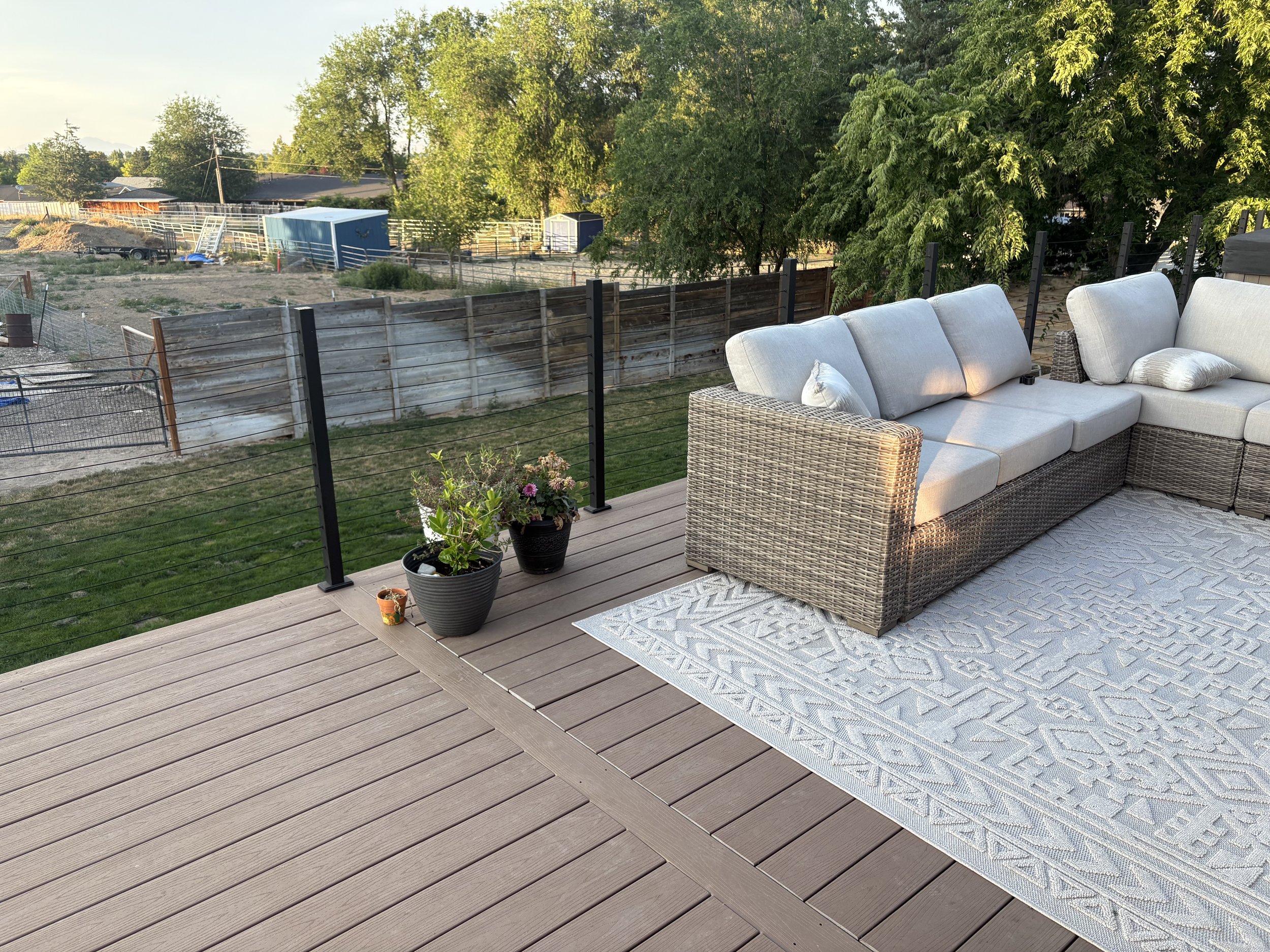 Outdoor patio with a wicker sectional sofa, a patterned white rug, two potted plants, and a view of a fenced backyard with trees and storage sheds.