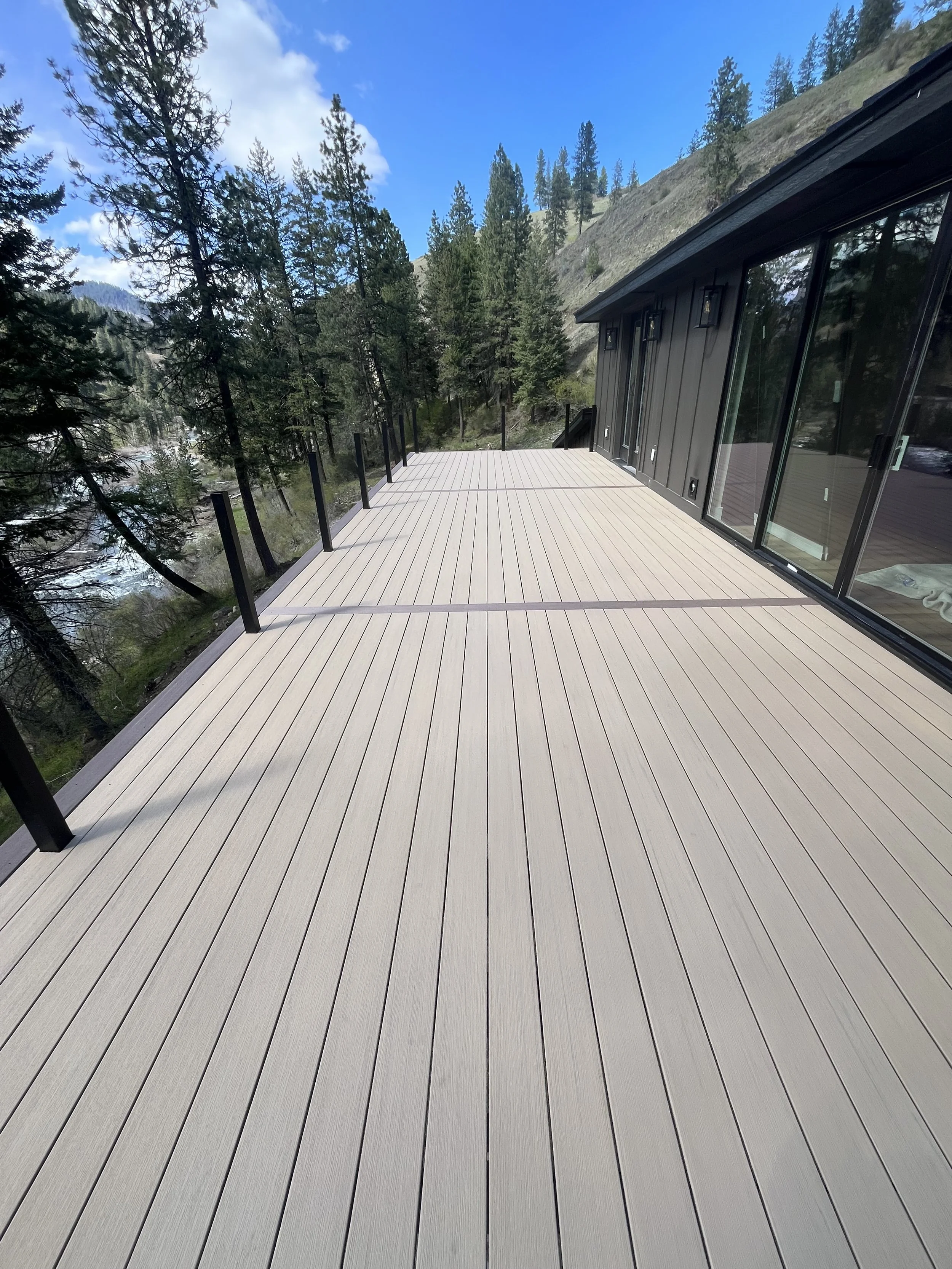 A spacious outdoor deck with light-colored wood planks, black metal railing posts, adjacent to a modern house with large glass sliding doors, overlooking a forested mountain landscape under a partly cloudy sky.