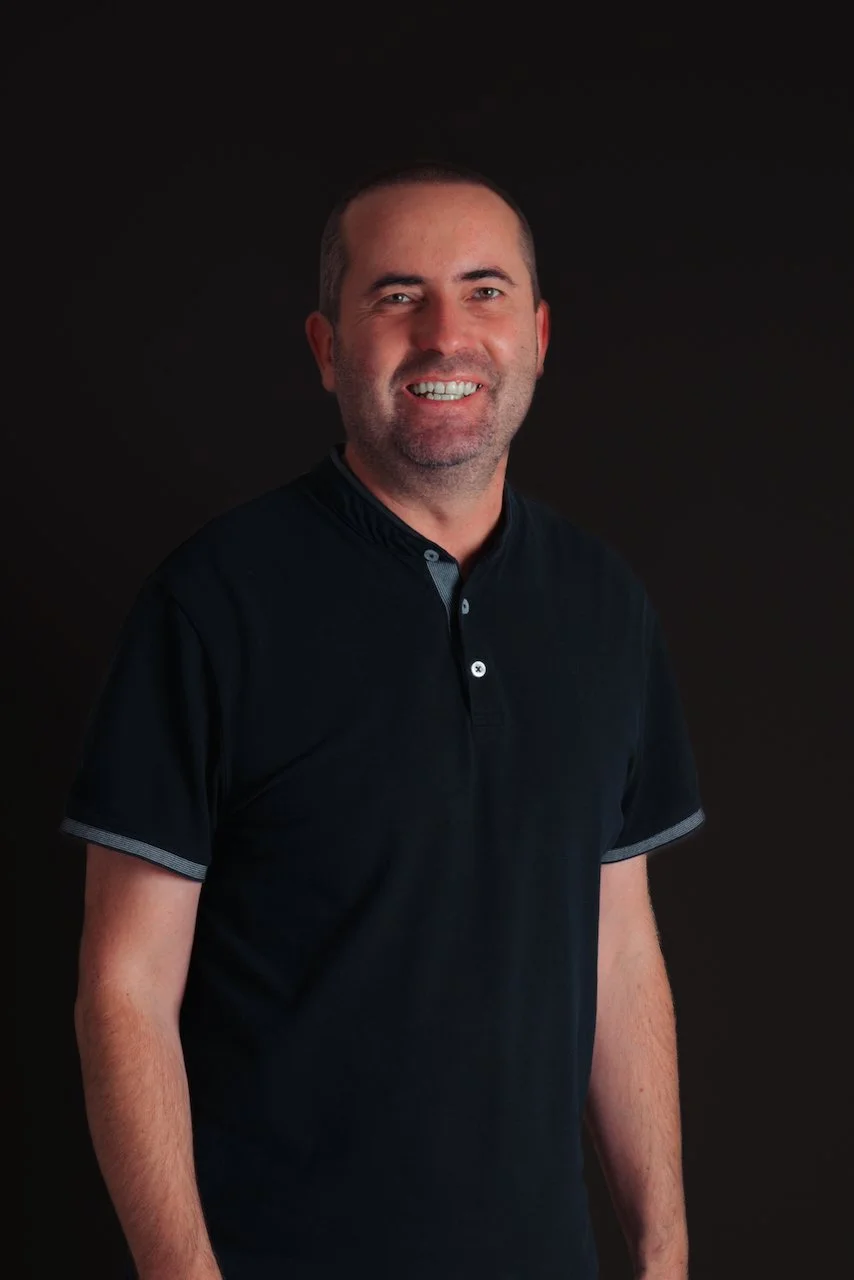 Un homme souriant portant un polo noir contre un fond noir.