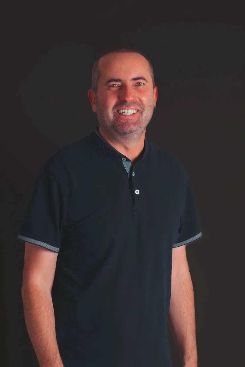 Un homme souriant portant une chemise noire, posé devant un fond sombre.