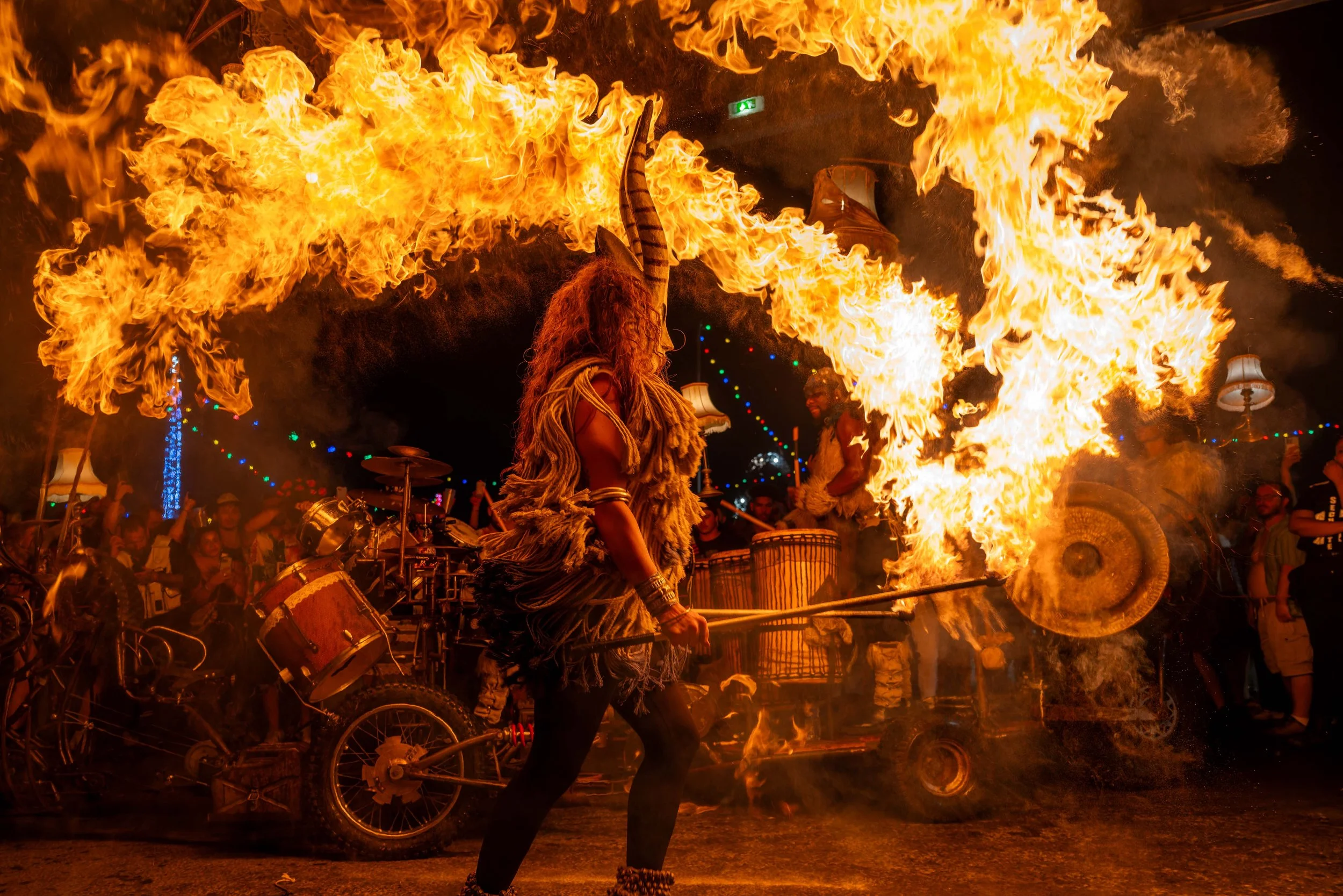 Un spectacle de feu avec des personnes en costumes traditionnels en arrière-plan, dans une ambiance festive avec des lumières colorées.