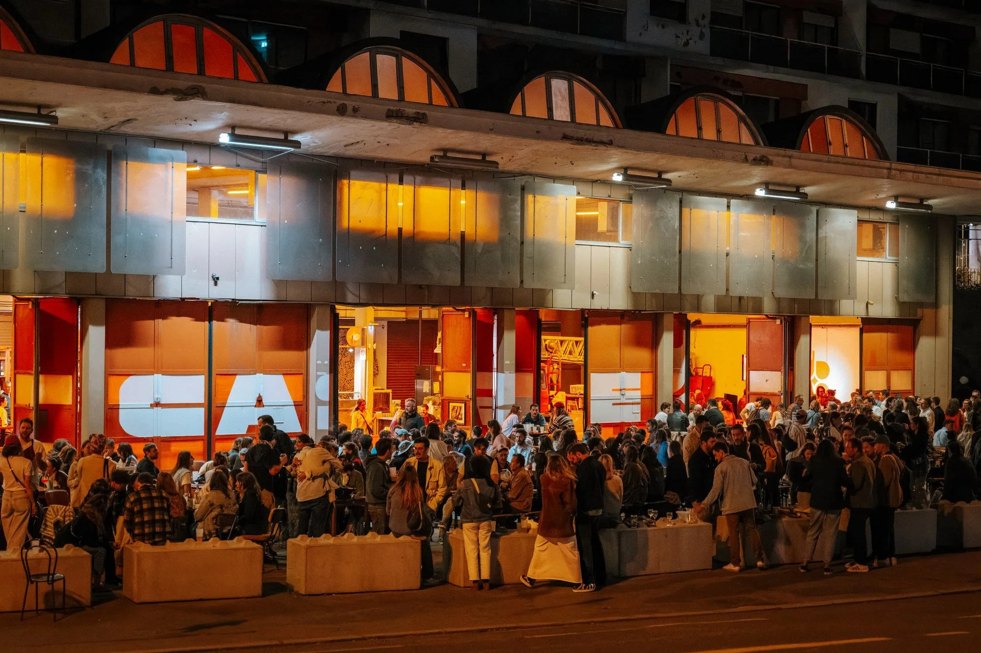 Une scène urbaine nocturne avec un grand nombre de personnes rassemblées devant un bâtiment illuminé aux couleurs chaudes, avec des fenêtres orange et un mobilier en bois.