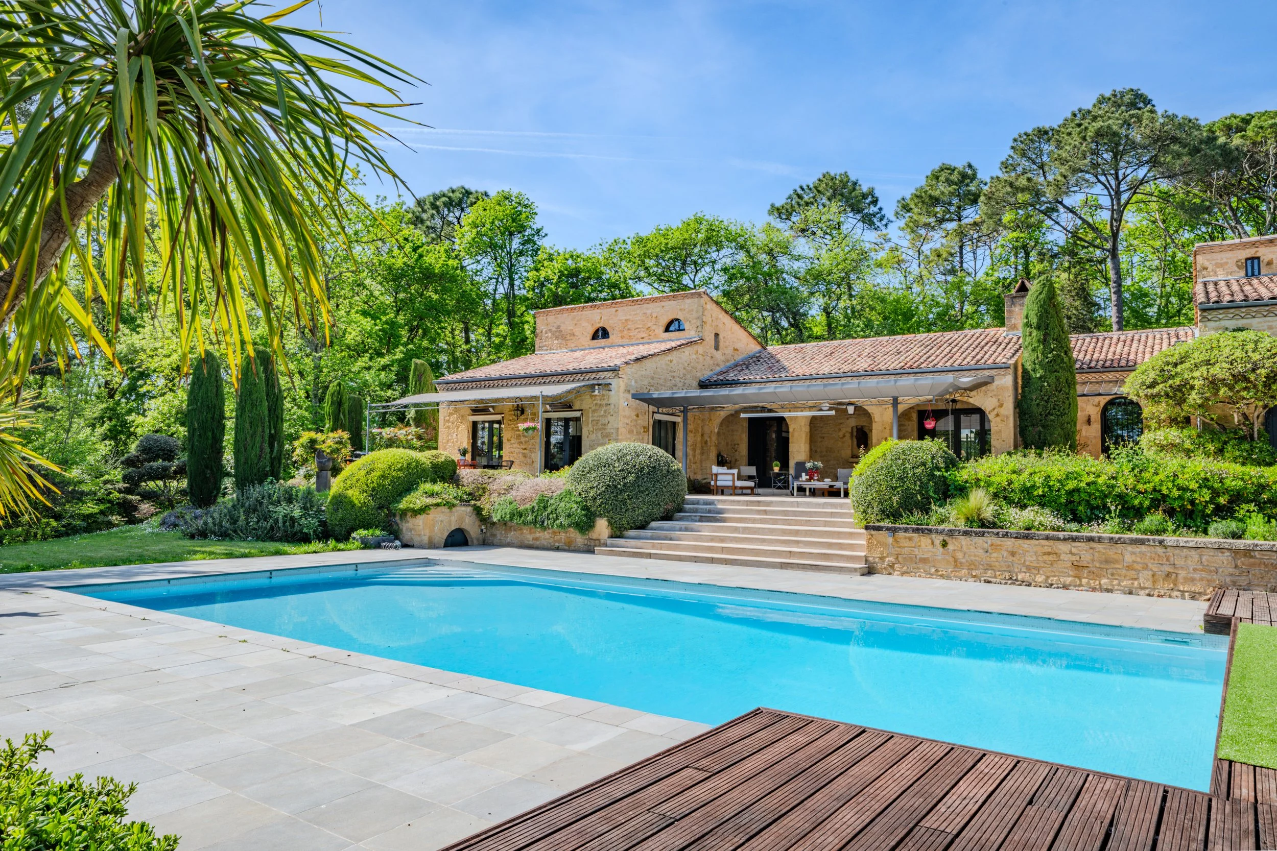Maison en pierre avec terrasse, jardin bien entretenu, piscine, et environnement verdoyant sous un ciel bleu.