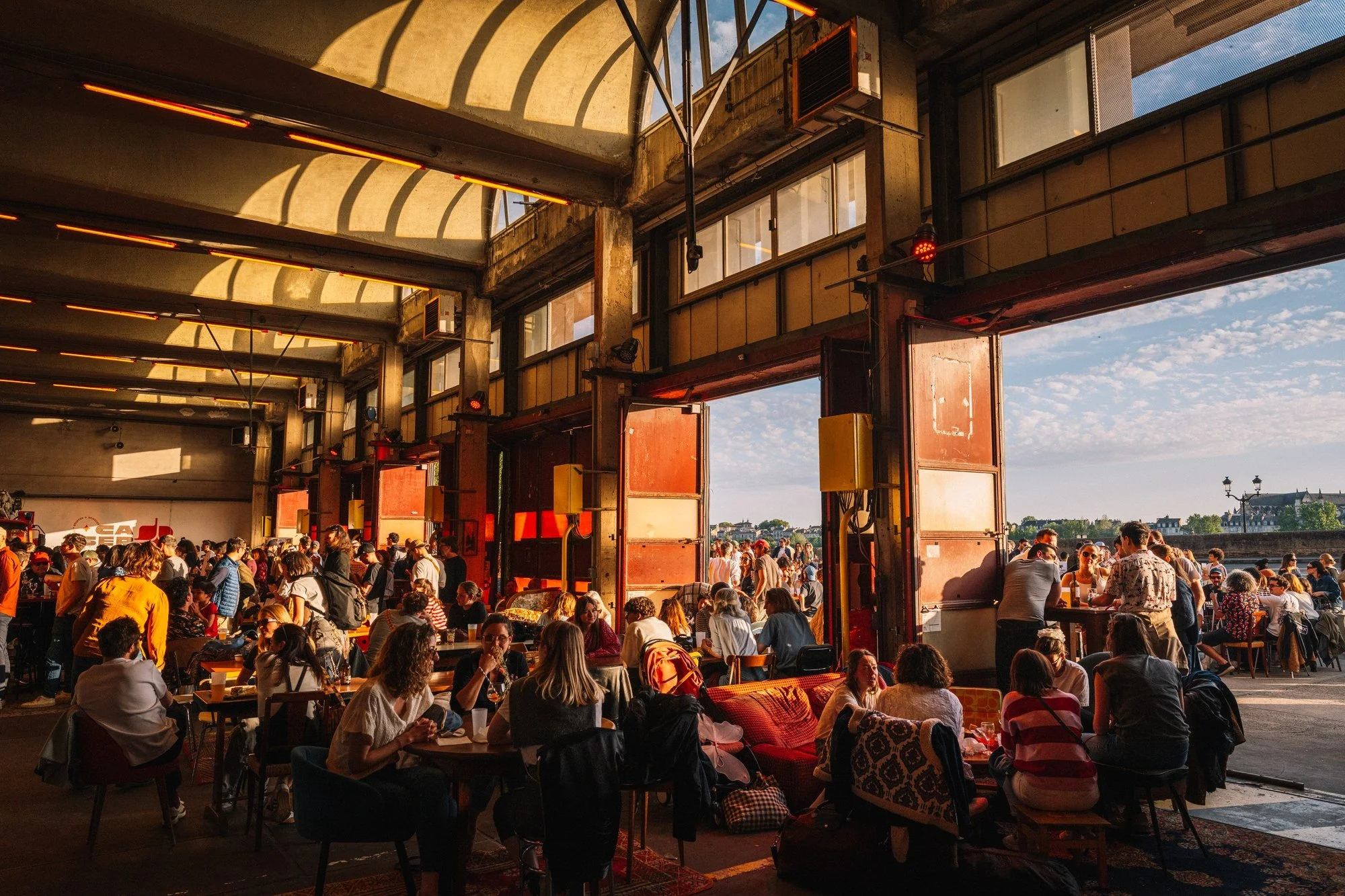 Une grande salle ouverte où de nombreuses personnes sont assises à des tables en train de manger ou de socialiser. La lumière naturelle pénètre par de grandes fenêtres et une ouverture en toiture, créant des ombres et une ambiance chaleureuse.