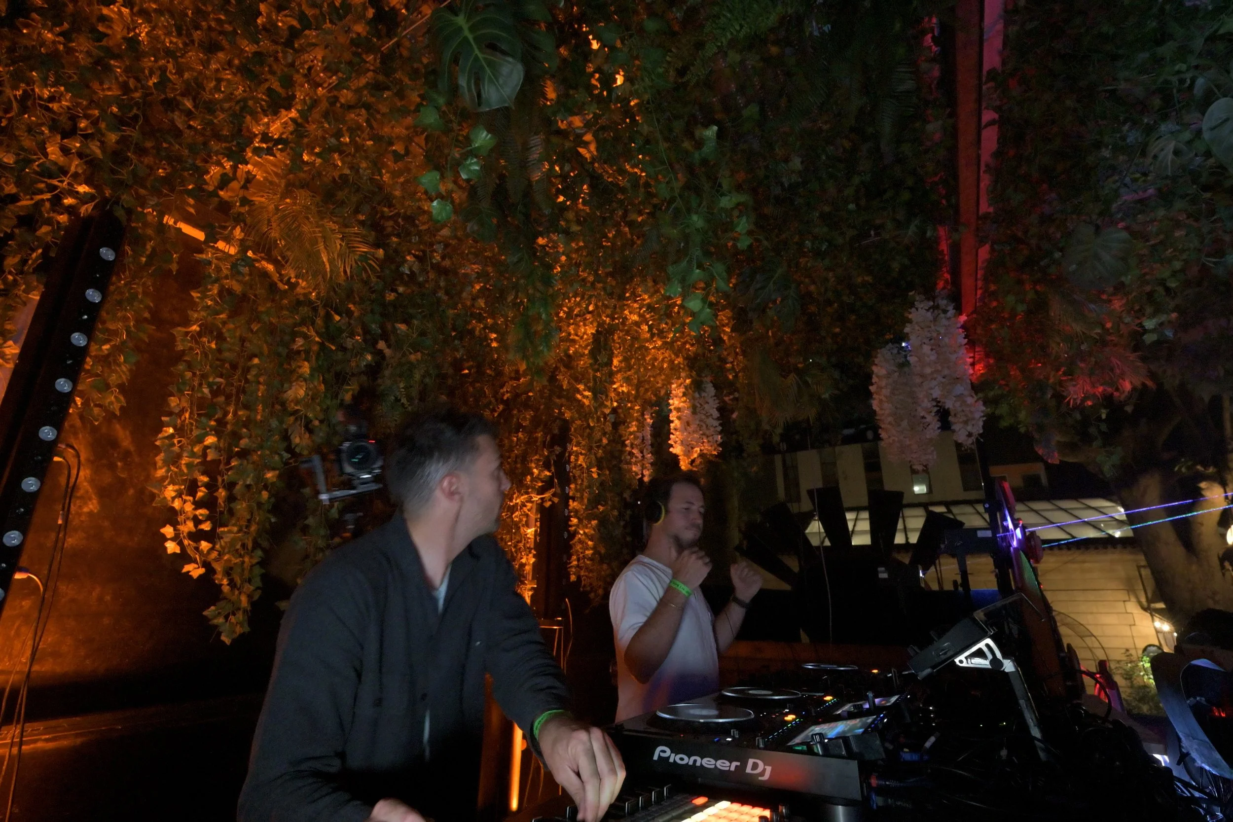 Deux DJ jouent de la musique sous un feuillage dense, avec un éclairage chaud orange et quelques touches de lumière colorée. Ils portent des écouteurs, et ils sont concentrés sur leurs appareils de DJ.