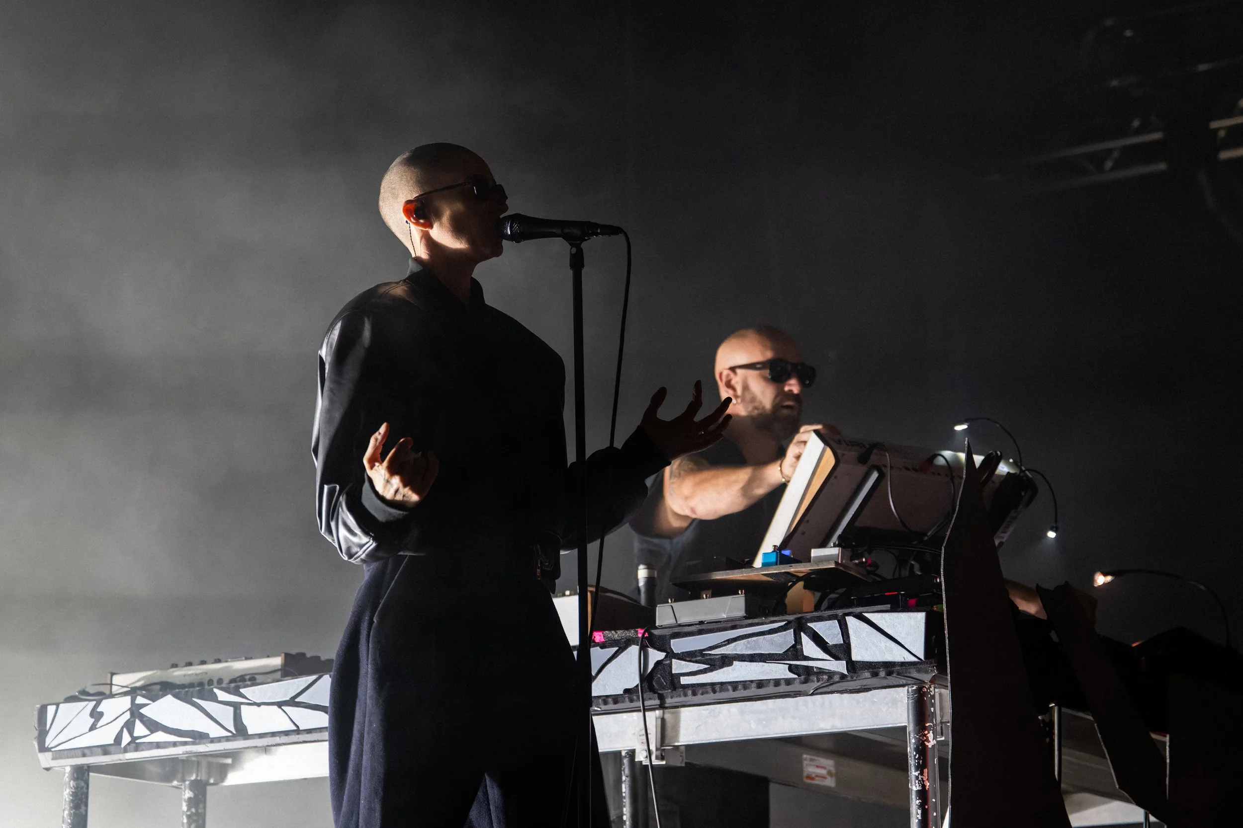 Deux musiciens, un homme et une femme, jouent sur scène avec des synthétiseurs et des microphones, dans une atmosphère sombre et brumeuse.
