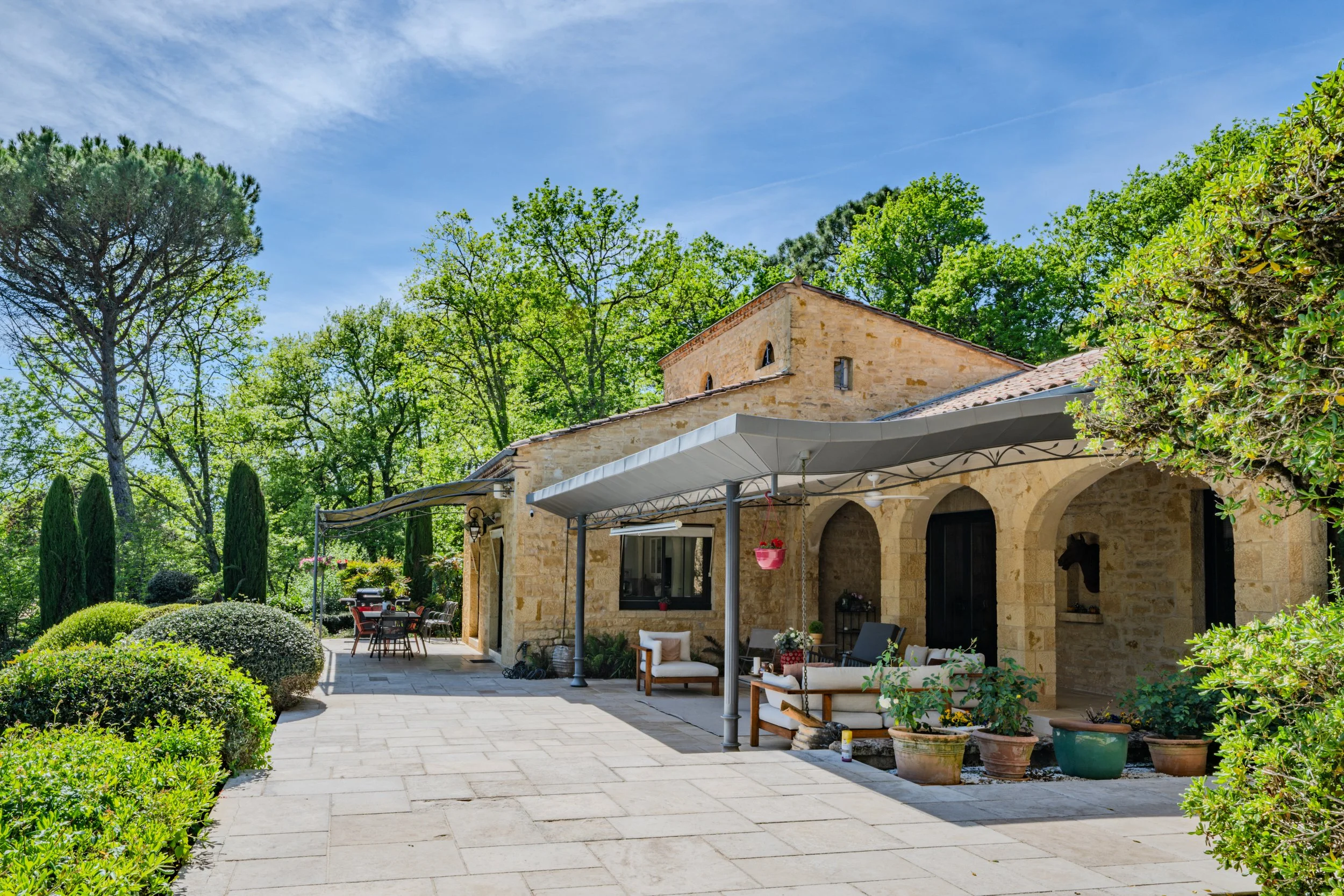 Une maison en pierre avec un porche, un jardin bien aménagé, des tables et des chaises en extérieur, entourée de végétation verdoyante et d'un ciel bleu clair.