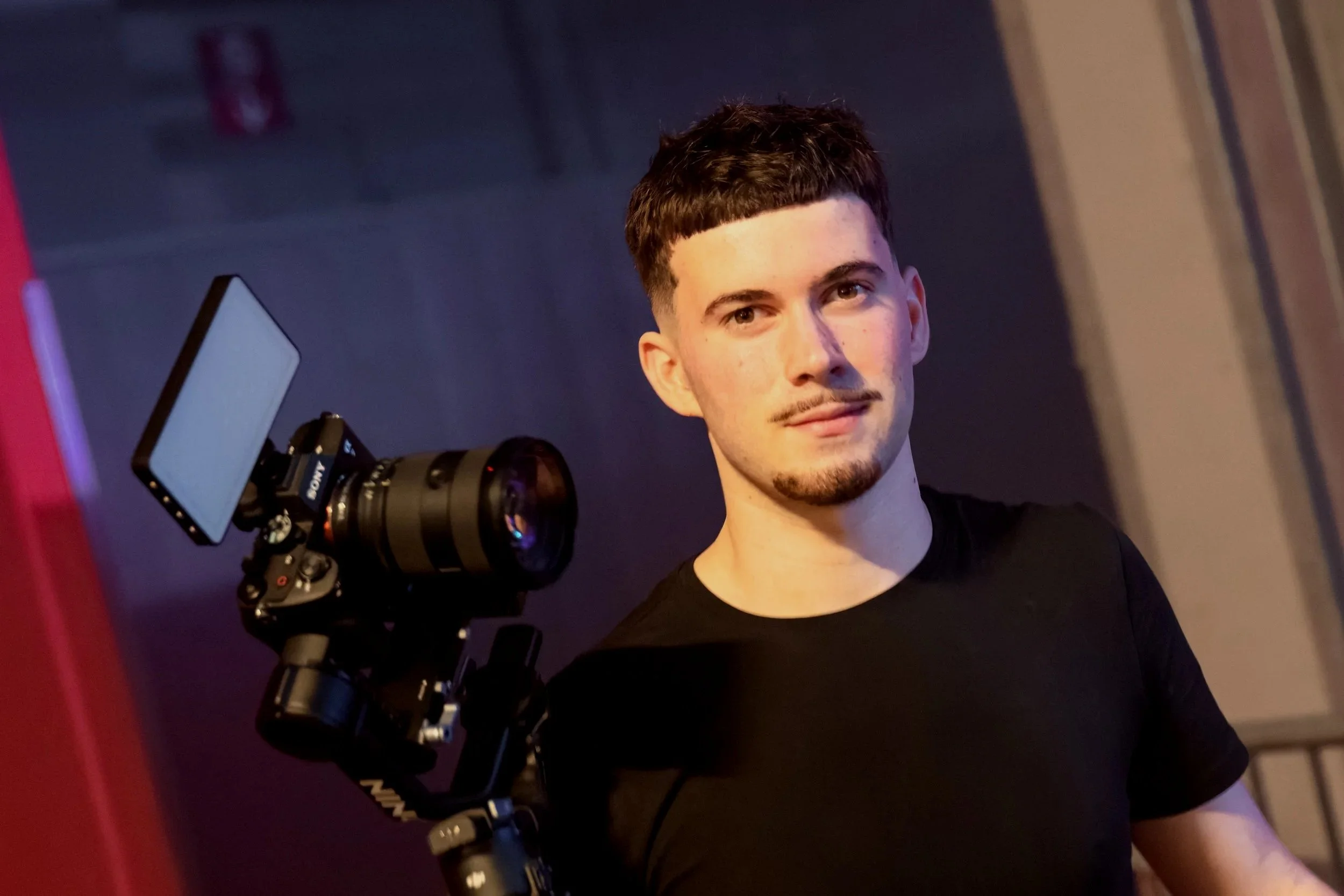 Jeune homme photographié à côté d'un appareil photo professionnel, en intérieur, avec un fond sombre.