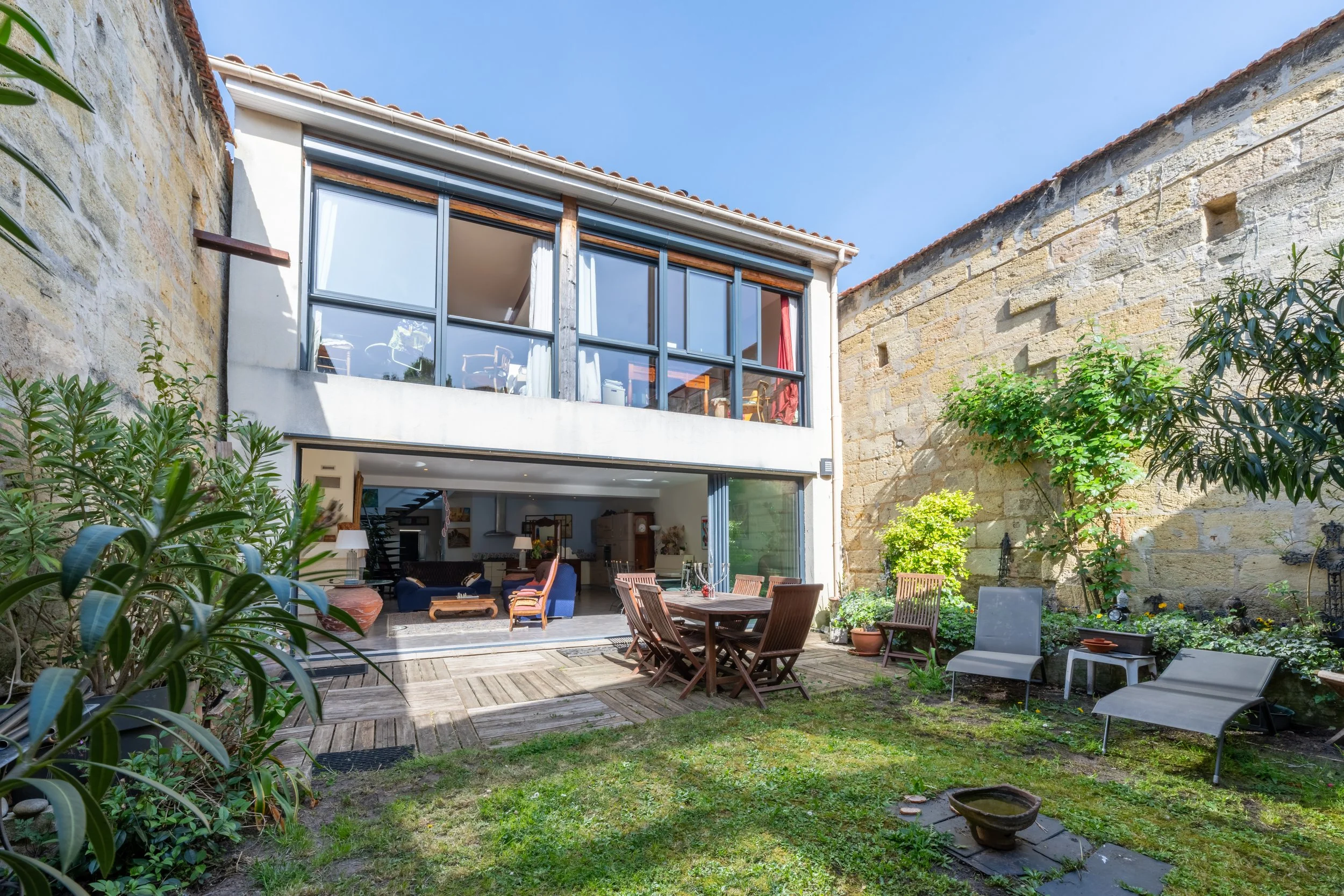 Jardin arrière avec fauteuils, table, plusieurs chaises en bois, plantes, mur en pierre et maison à deux étages avec balcon en verre.