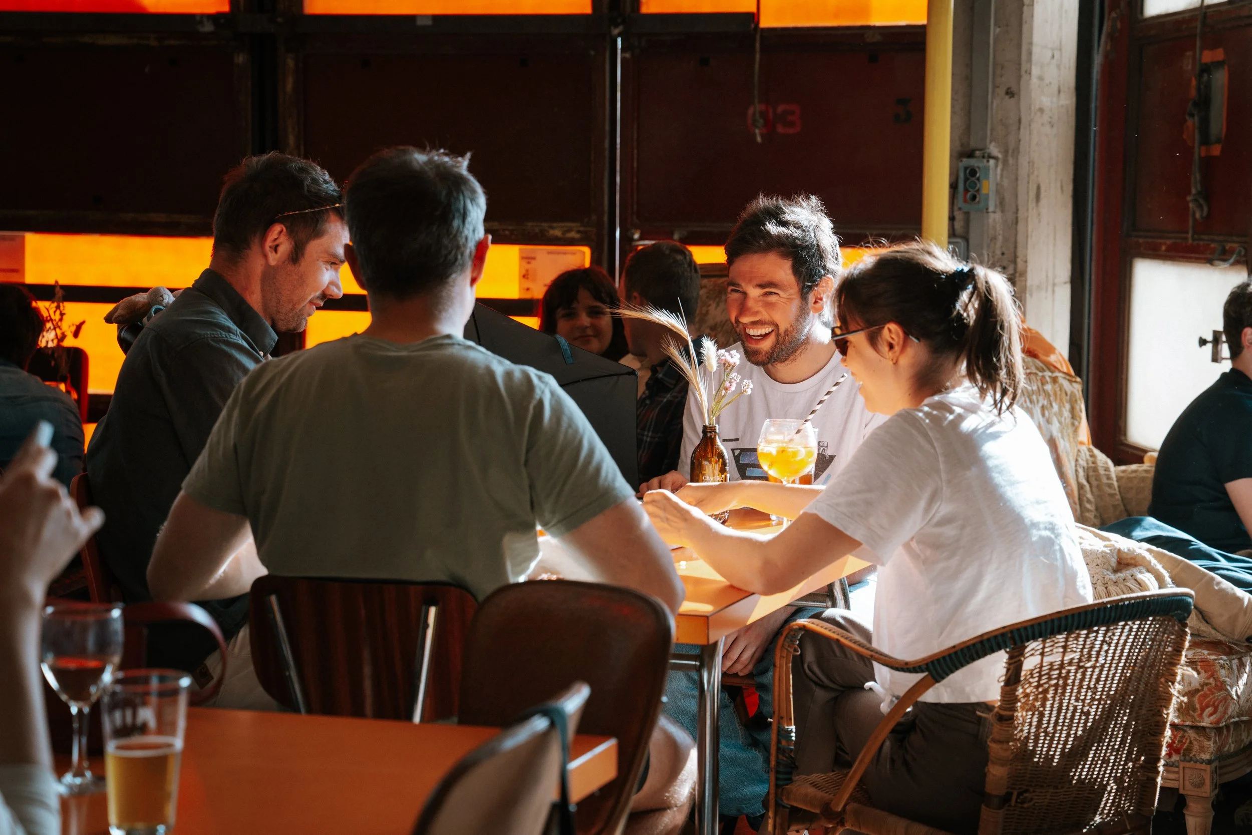 Groupe de personnes assises autour d'une table en train de discuter et de rire dans un restaurant ou un café. Le décor est industriel avec des murs en béton et des panneaux de bois, éclairage chaud.