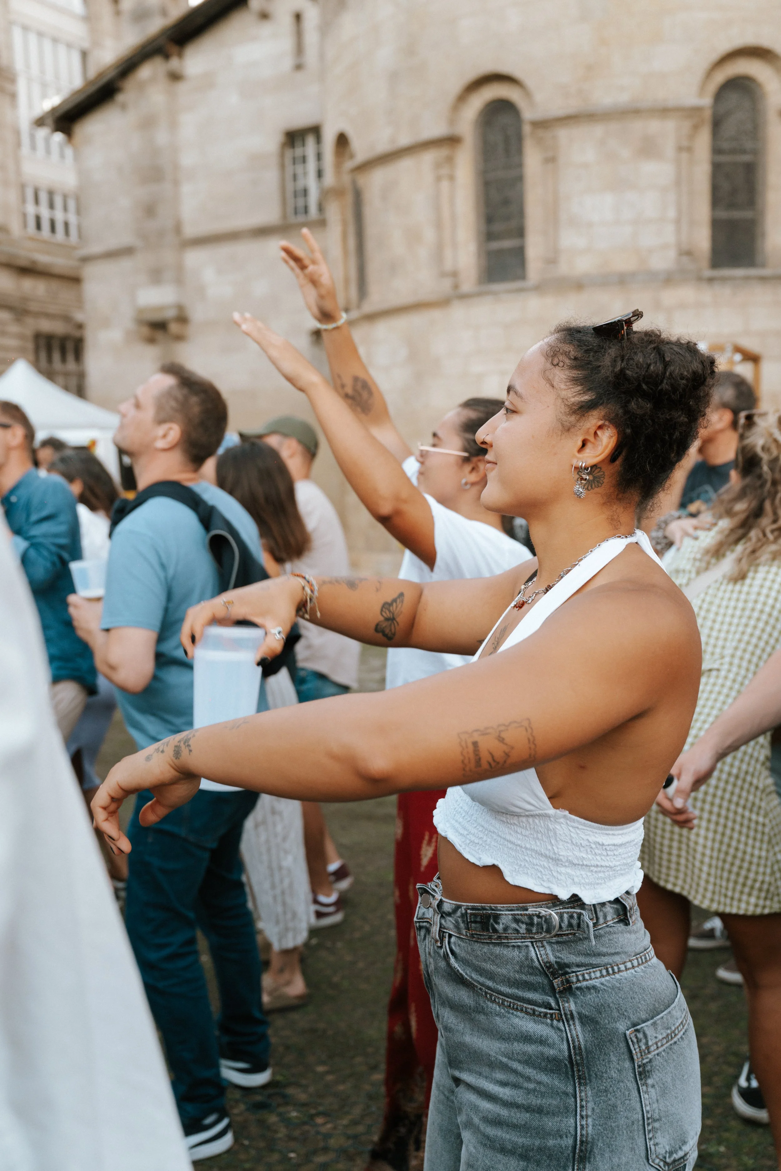Groupe de personnes assistantes à un événement en extérieur, parmi elles une femme avec des tatouages, des bijoux, un haut blanc et un jean, souriant, avec d'autres personnes devant un bâtiment en pierre.