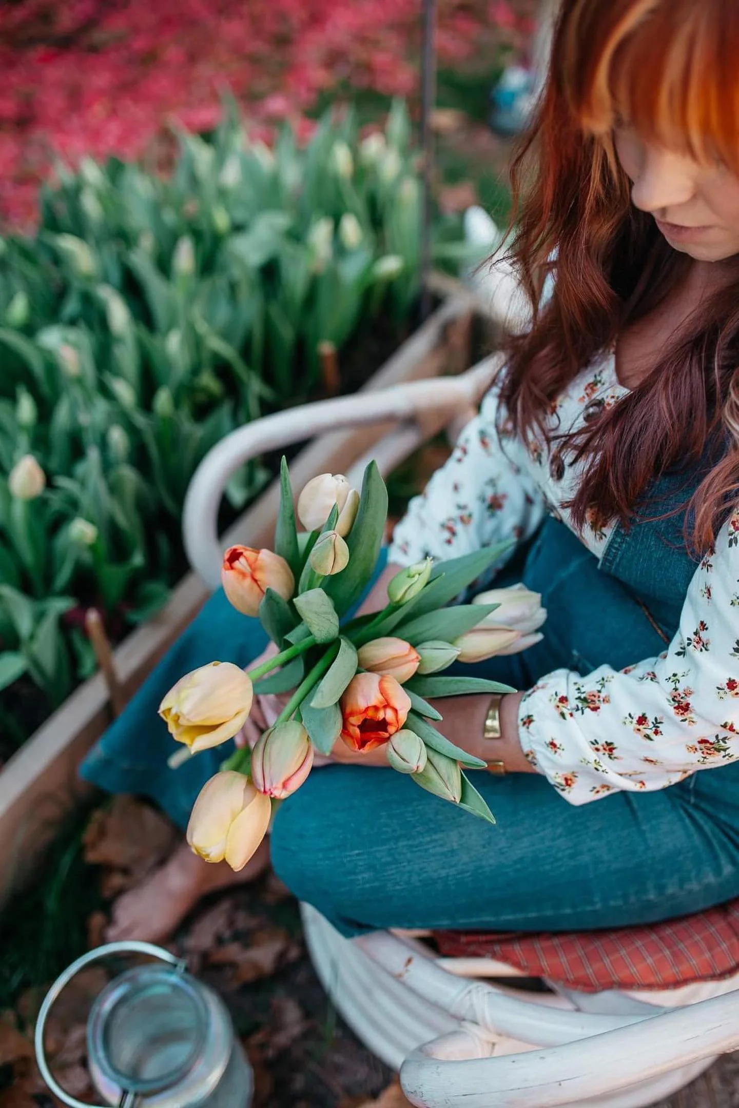 A woman with long red hair sitting on a white chair holds a bouquet of tulips while surrounded by tulip plants in a garden.
