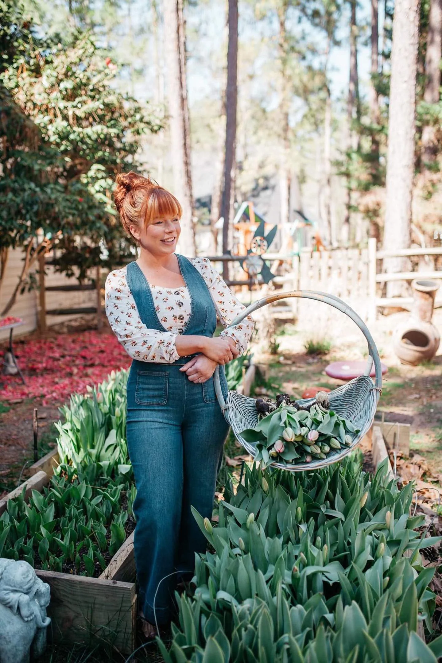 Woman with red hair, wearing floral blouse and denim overalls, standing in a garden with tulips, holding a basket of tulips.