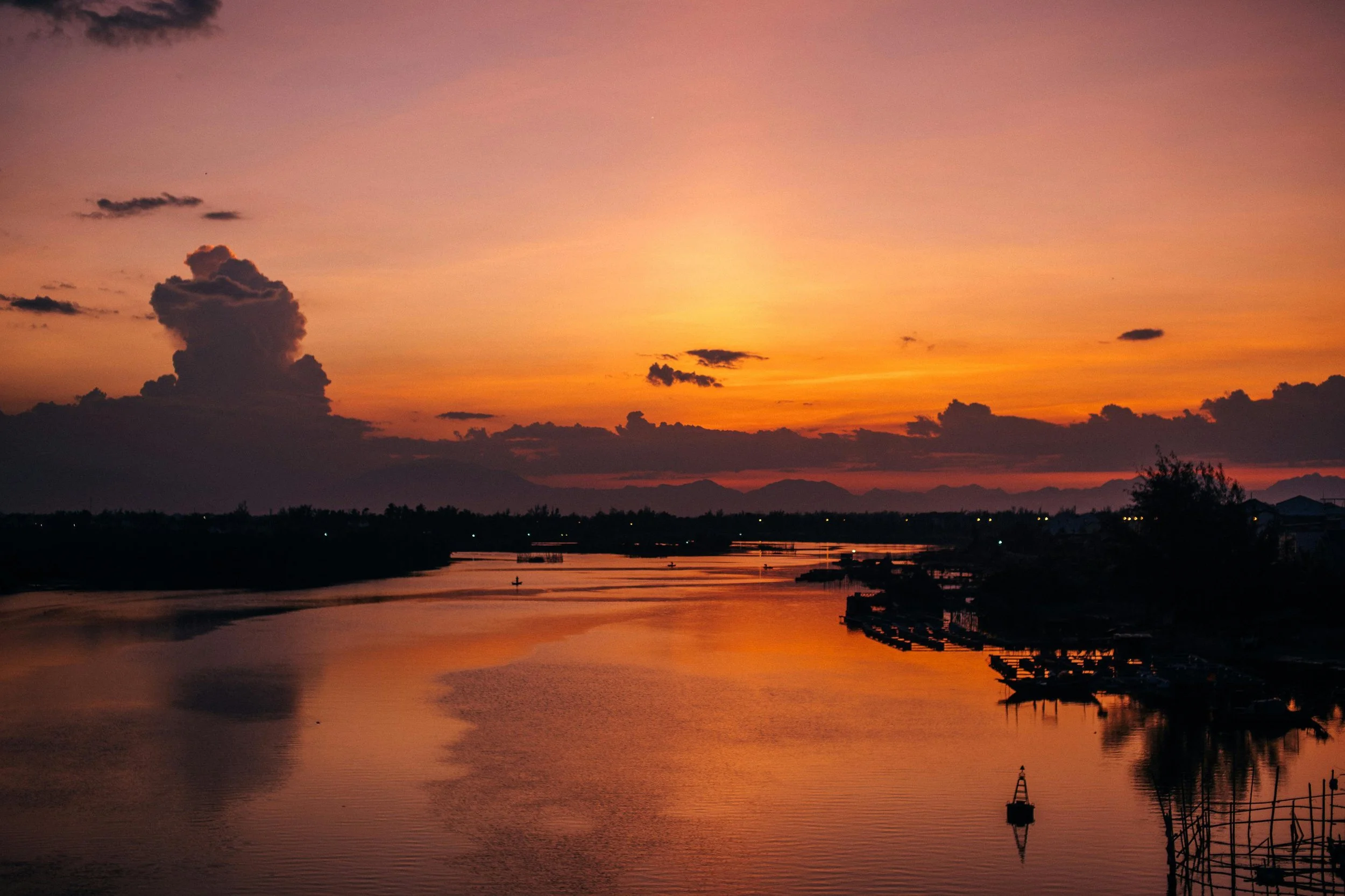 Sunset view over a river with boats and silhouettes of buildings and trees along the shoreline.