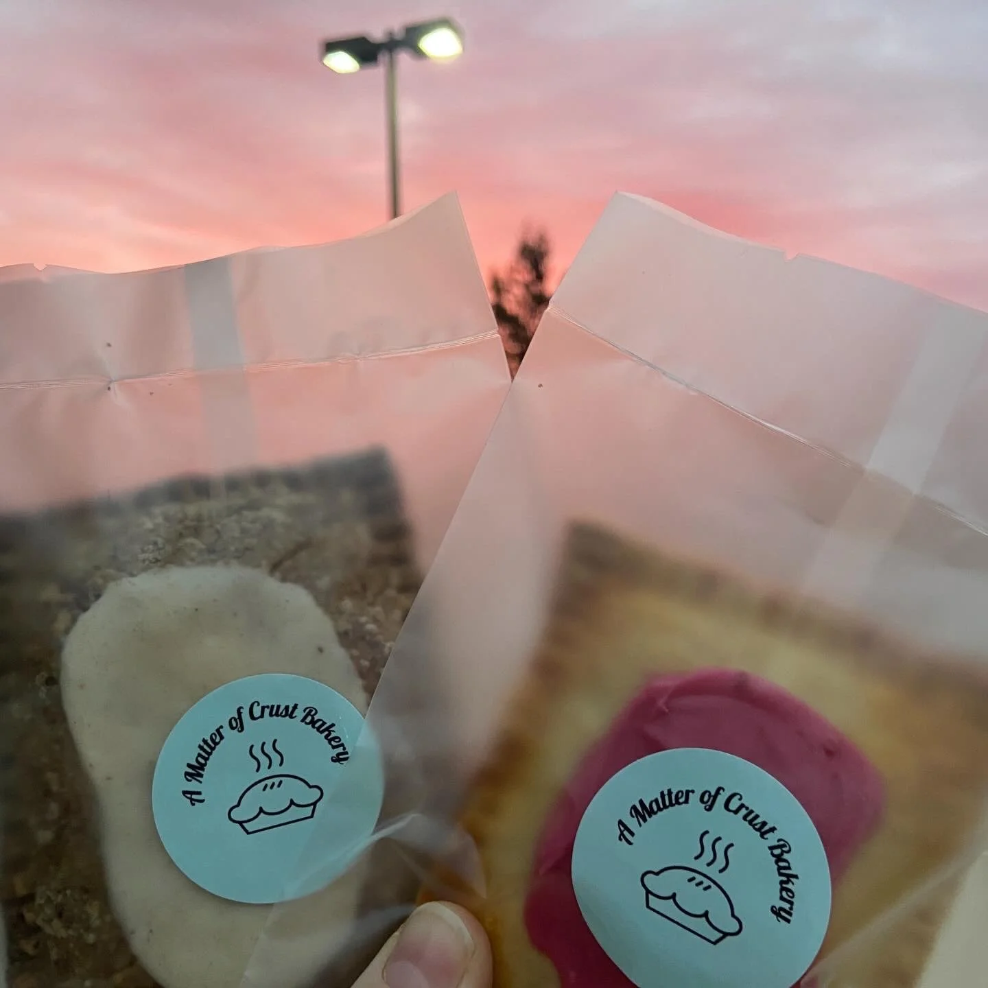 Two clear plastic bags with baked goods inside, one containing a brown rectangular pastry with icing and the other a rectangular pastry with pink icing, held up against a background of a pink and purple sunset sky and outdoor light post.