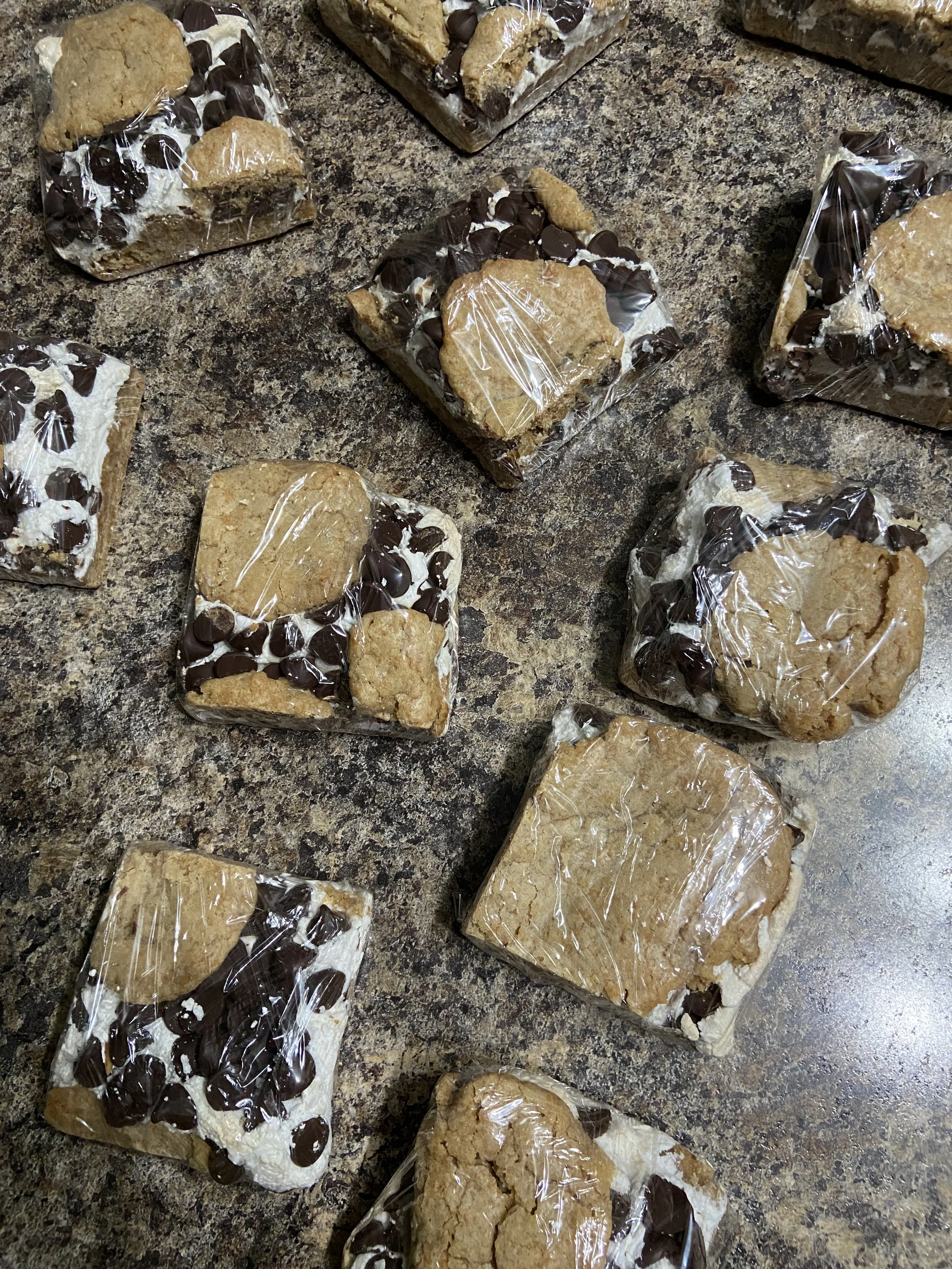 Multiple homemade cookies with chocolate chips, individually wrapped in plastic, placed on a speckled countertop.