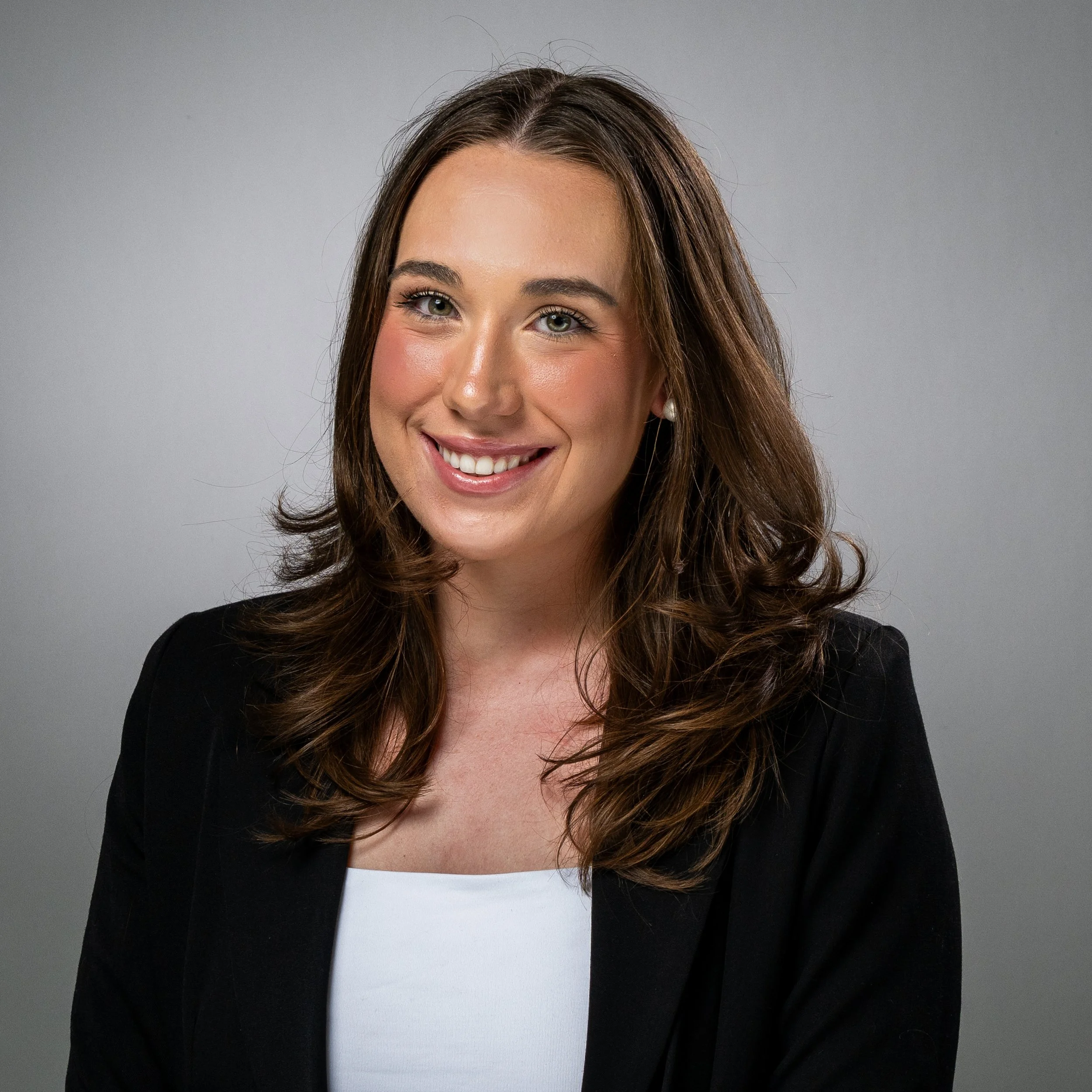 Headshot of a smiling woman with long wavy brown hair, wearing a black blazer over a white top, against a gray background.