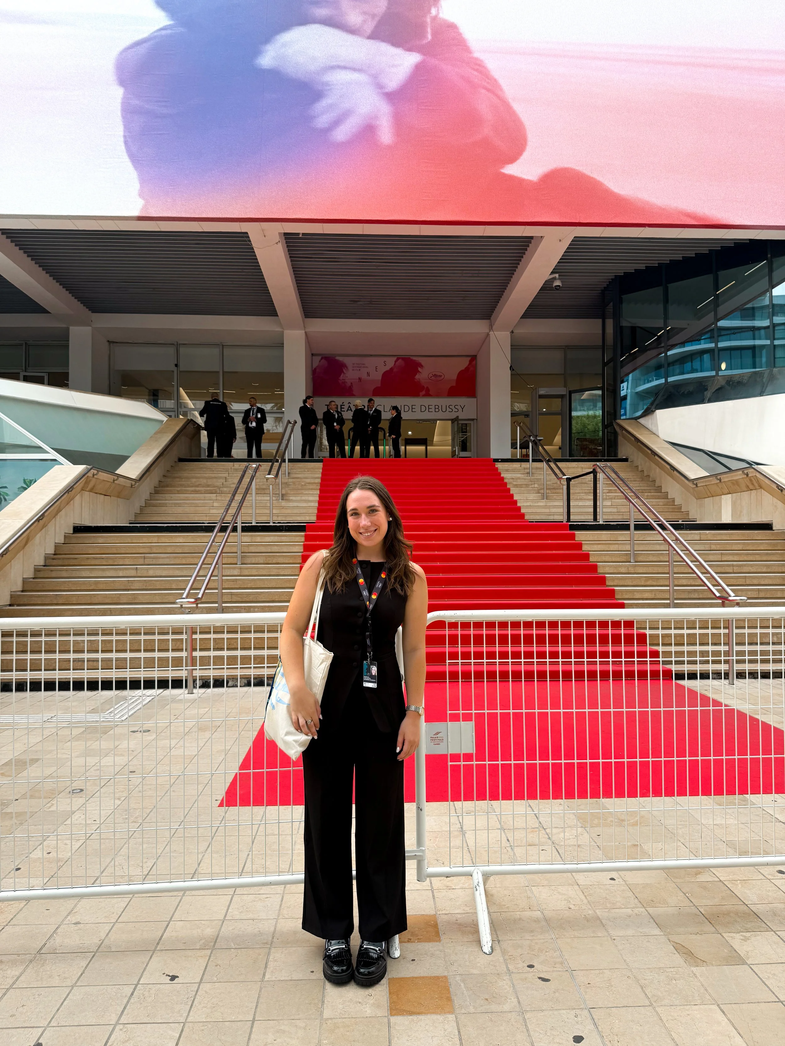A woman standing in front of a red carpet at the entrance of an event venue, with a large screen above displaying an image of two people hugging.