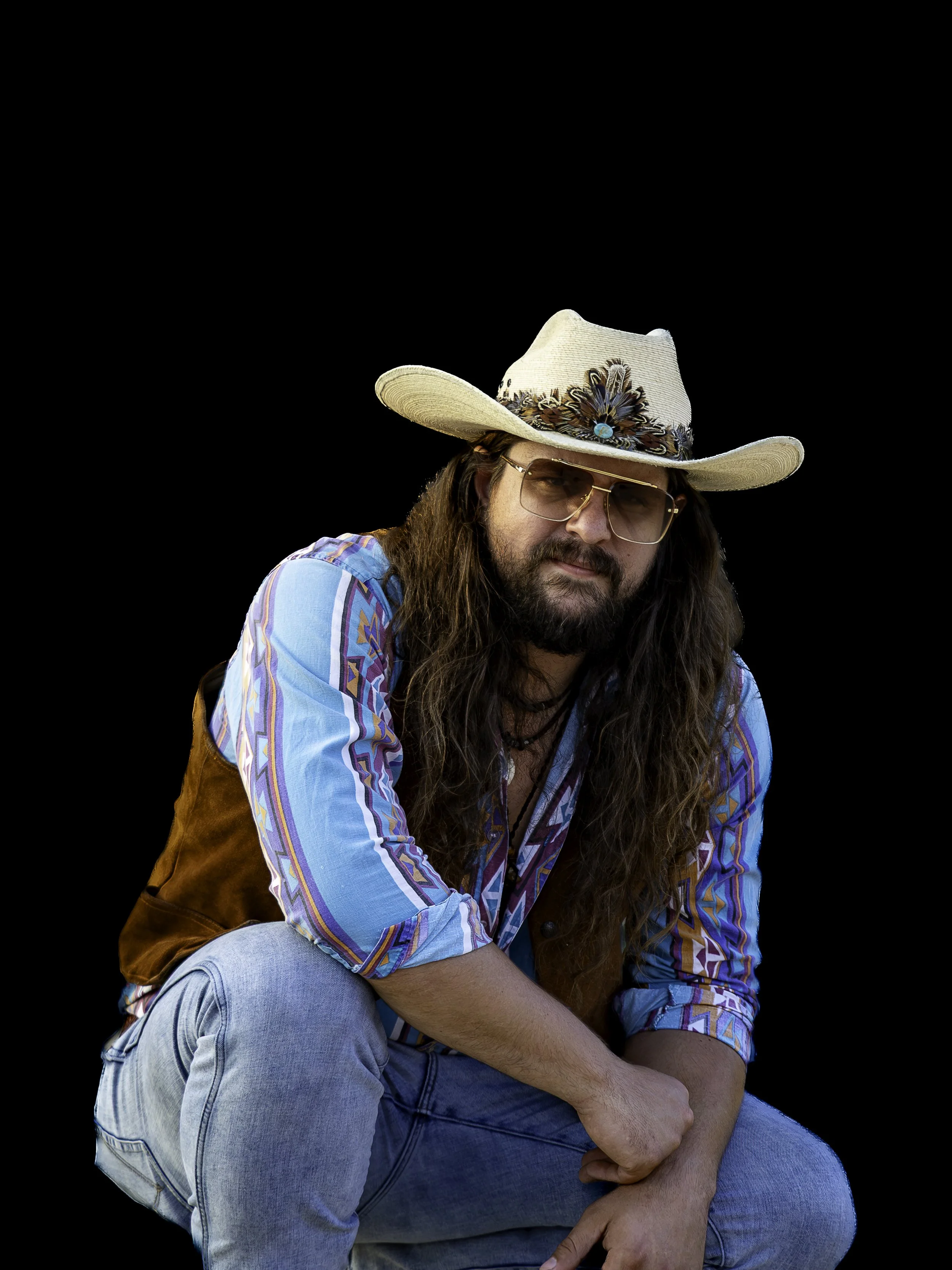 A man with long hair, a beard, wearing sunglasses, a cowboy hat decorated with feathers and stones, a colorful shirt, and a brown vest, sitting with his arms crossed against a black background.