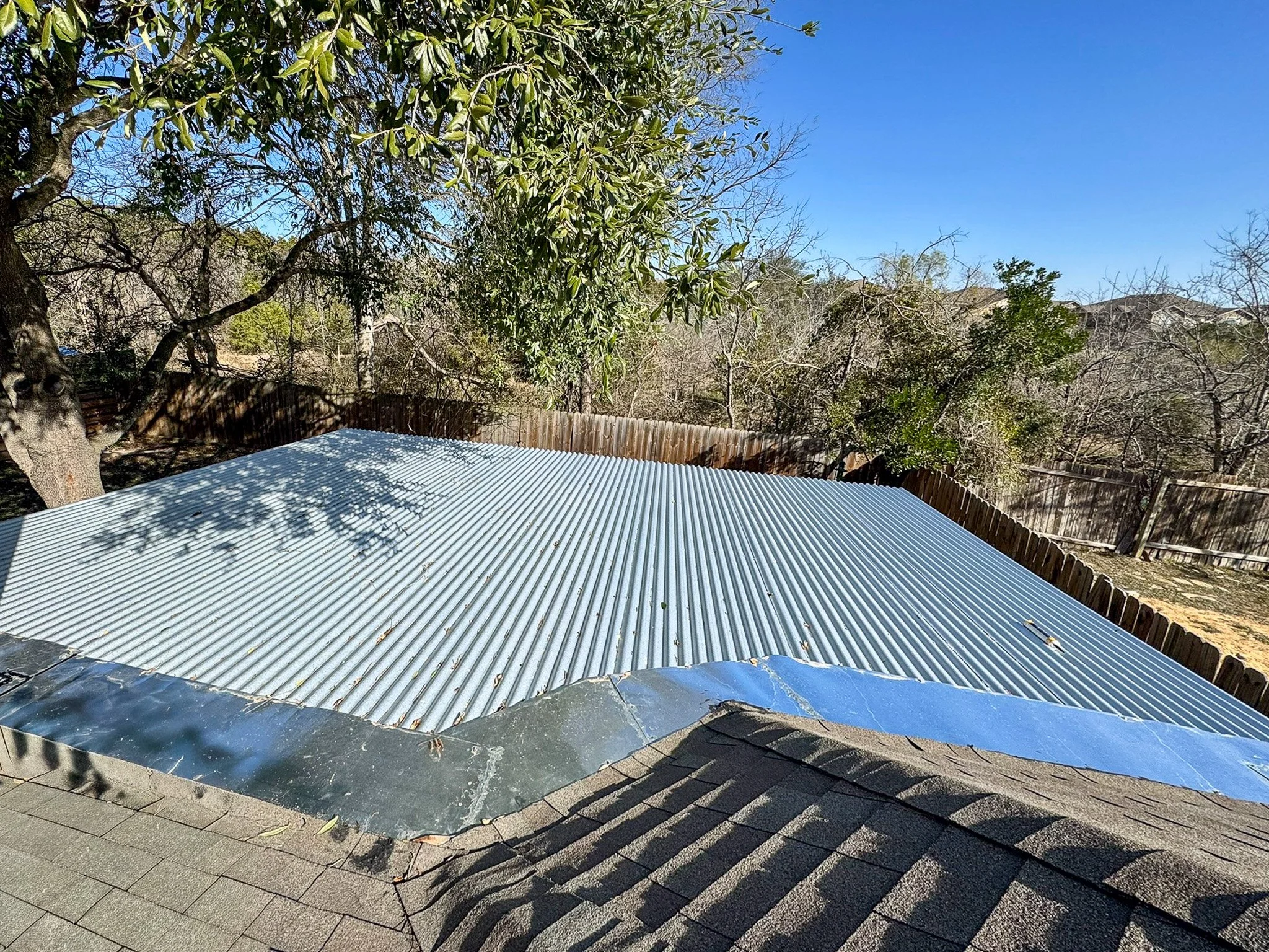 View of a backyard with a corrugated metal roof in the foreground, a wooden fence, and leafless and green trees against a clear blue sky.
