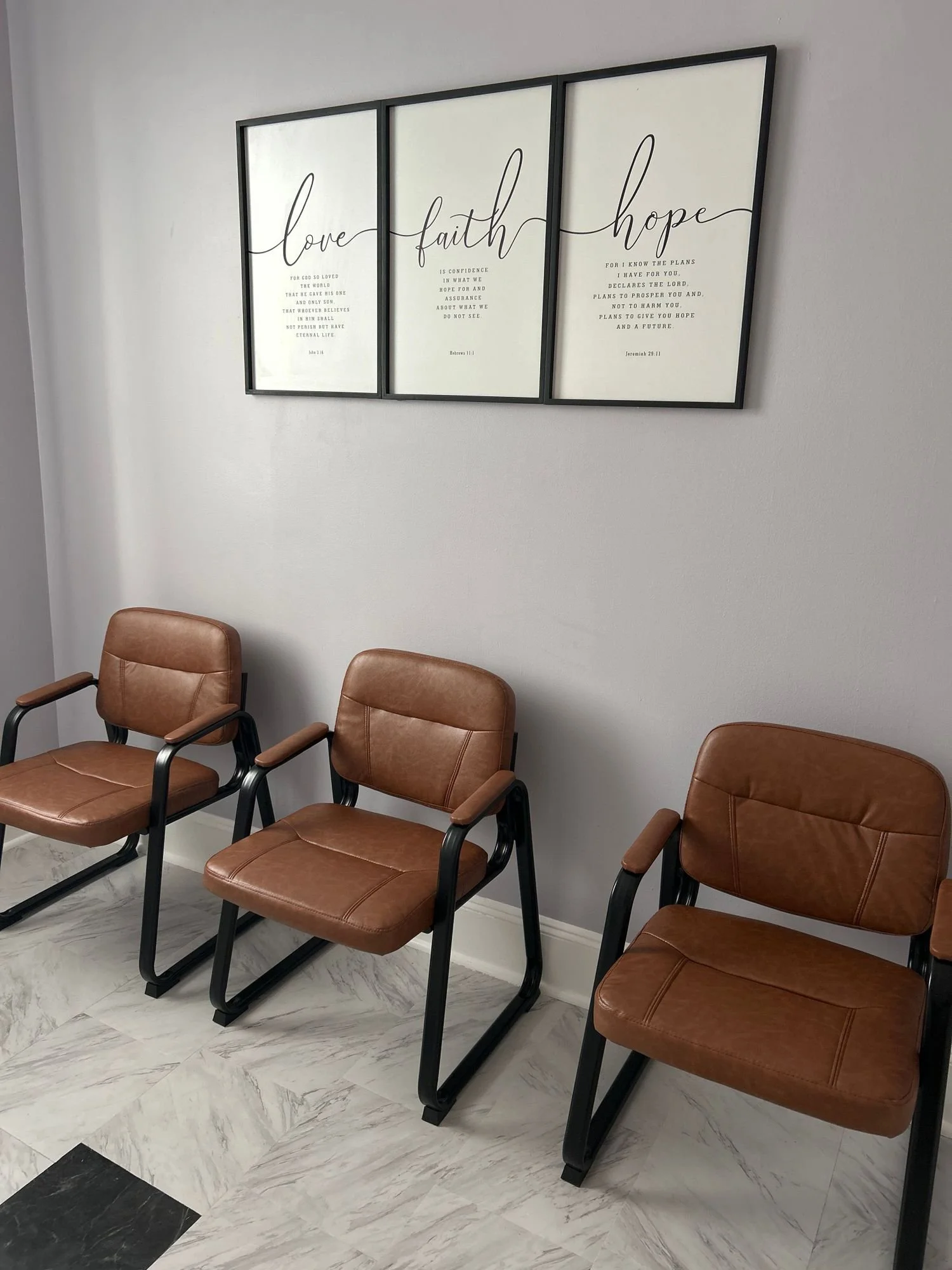 Three brown chairs with black metal frames arranged in a row in a waiting area at Stepping Stones Counseling Services next to a light gray wall with three framed posters containing inspirational words.