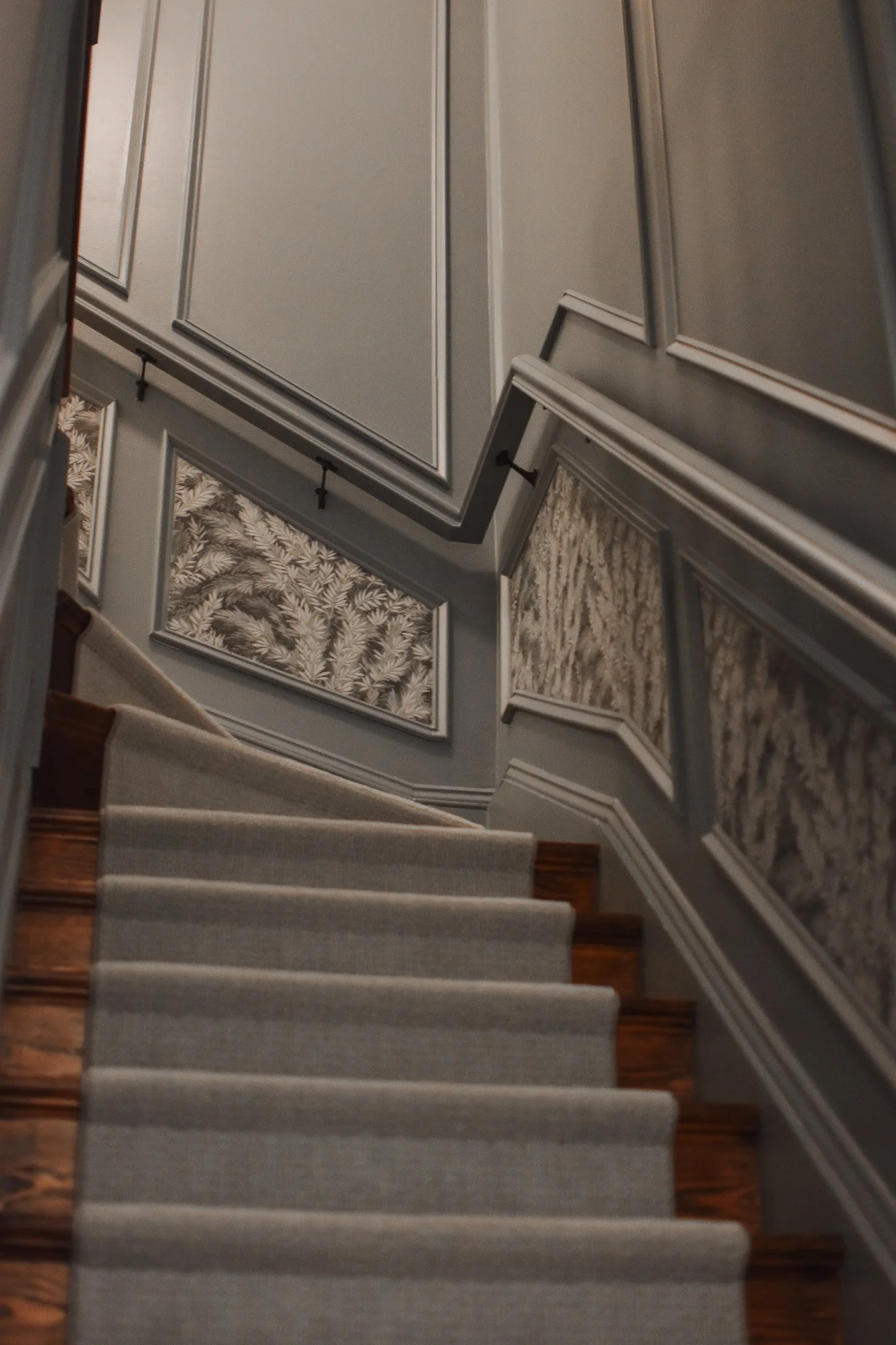 Indoor staircase with beige runner carpet, wooden stairs on the sides, and gray walls decorated with framed wallpaper panels featuring floral and botanical patterns.