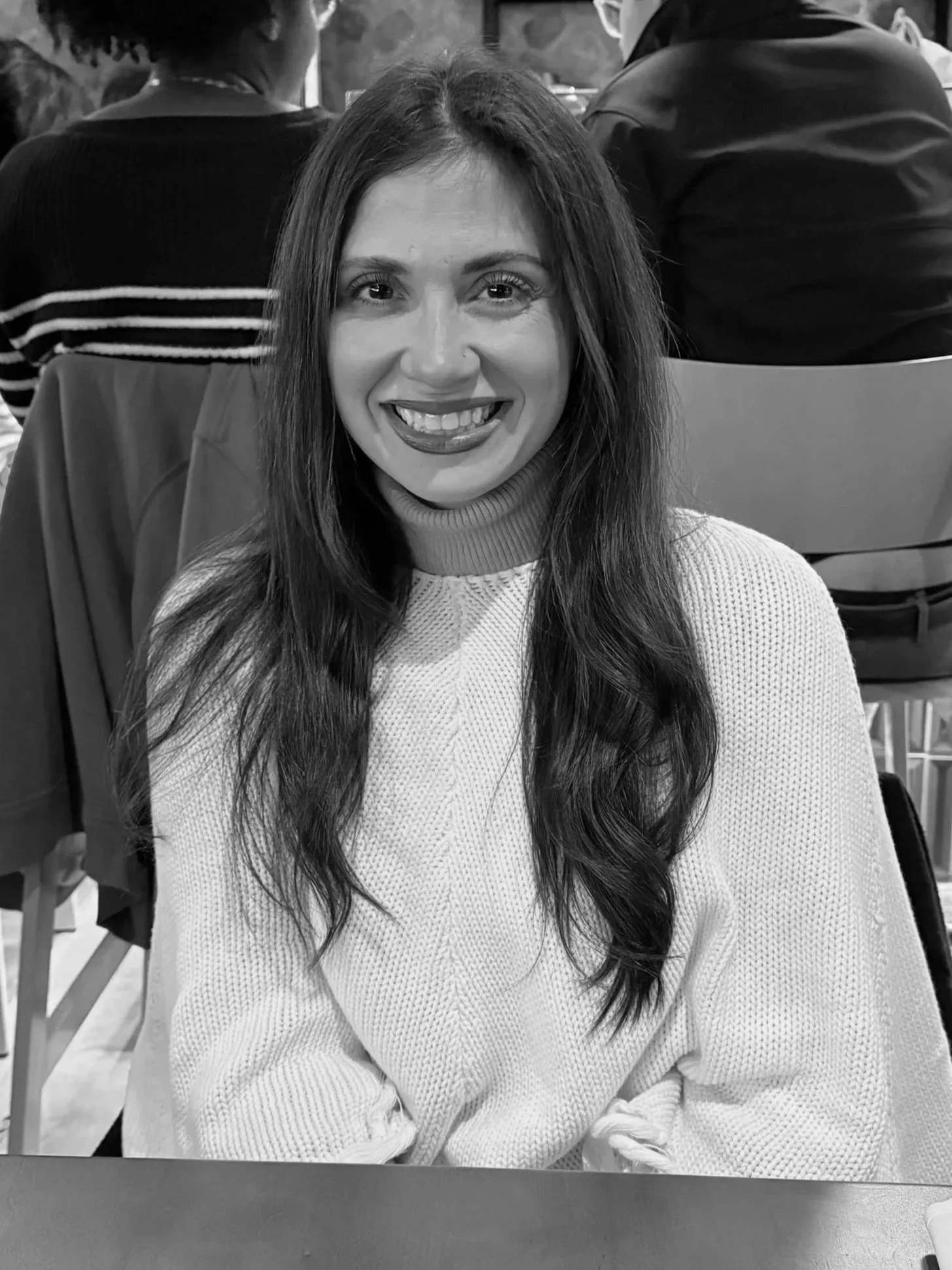 A woman with long dark hair smiling at the camera in a crowded indoor setting, wearing a light-colored sweater.
