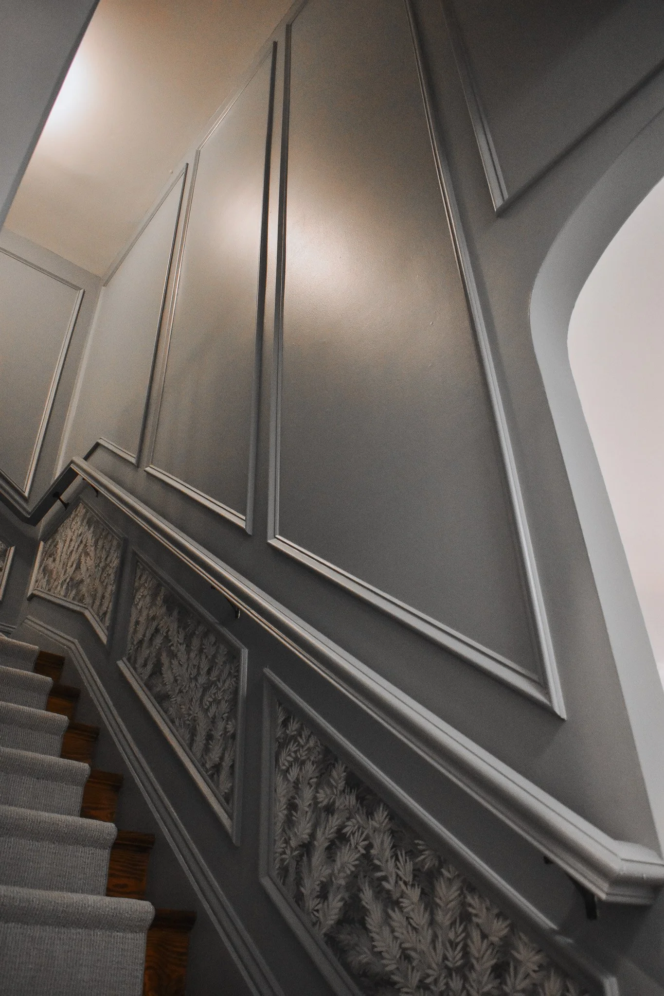 Interior view of a staircase with ornate wall paneling and molding, seen from below, with a high ceiling and a neutral-colored wall.