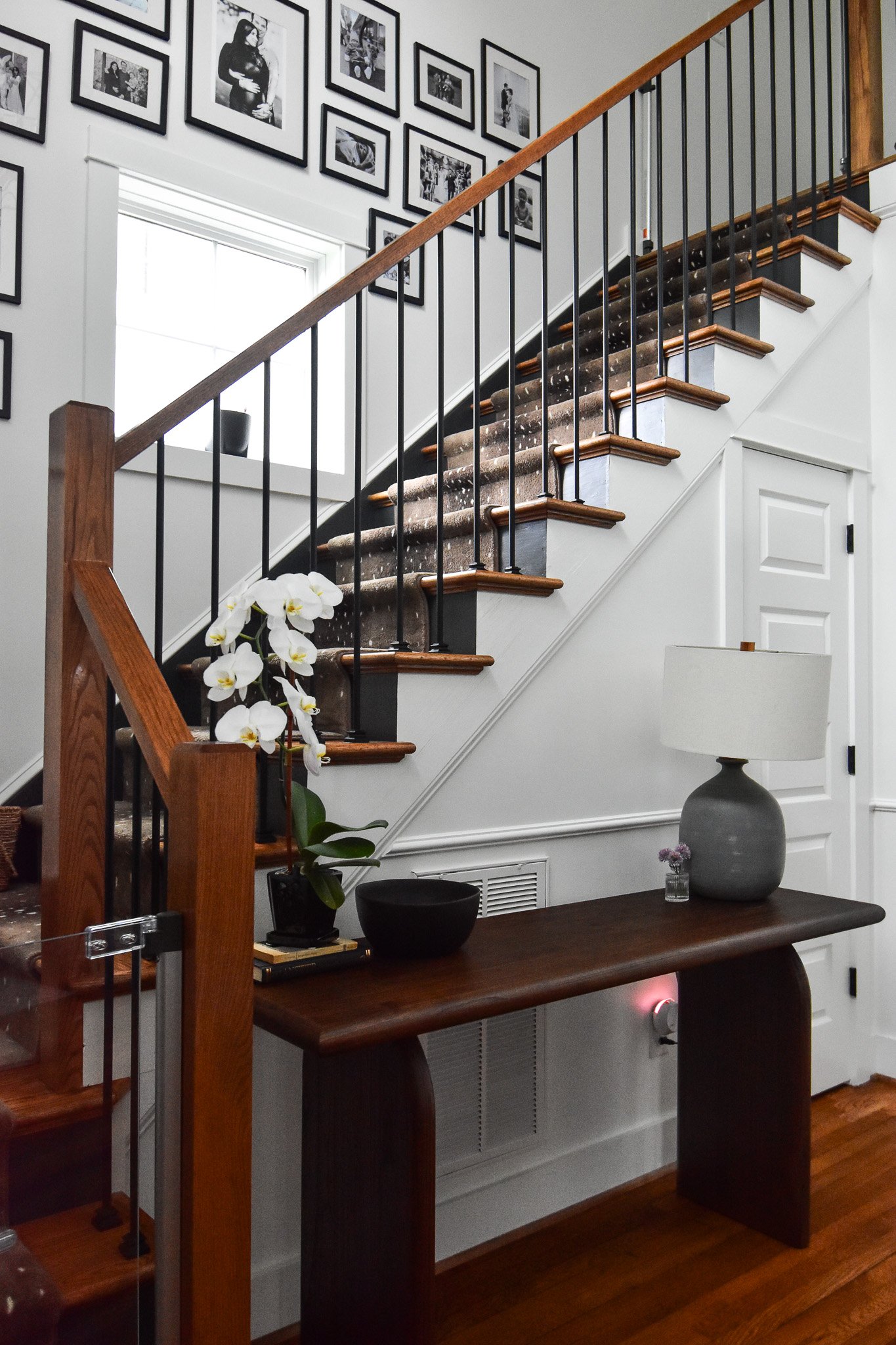 Interior view of a house featuring a staircase with a dark carpet runner, wall gallery of black and white photos, and a wooden console table with a white orchid, a gray lamp, and decorative items.
