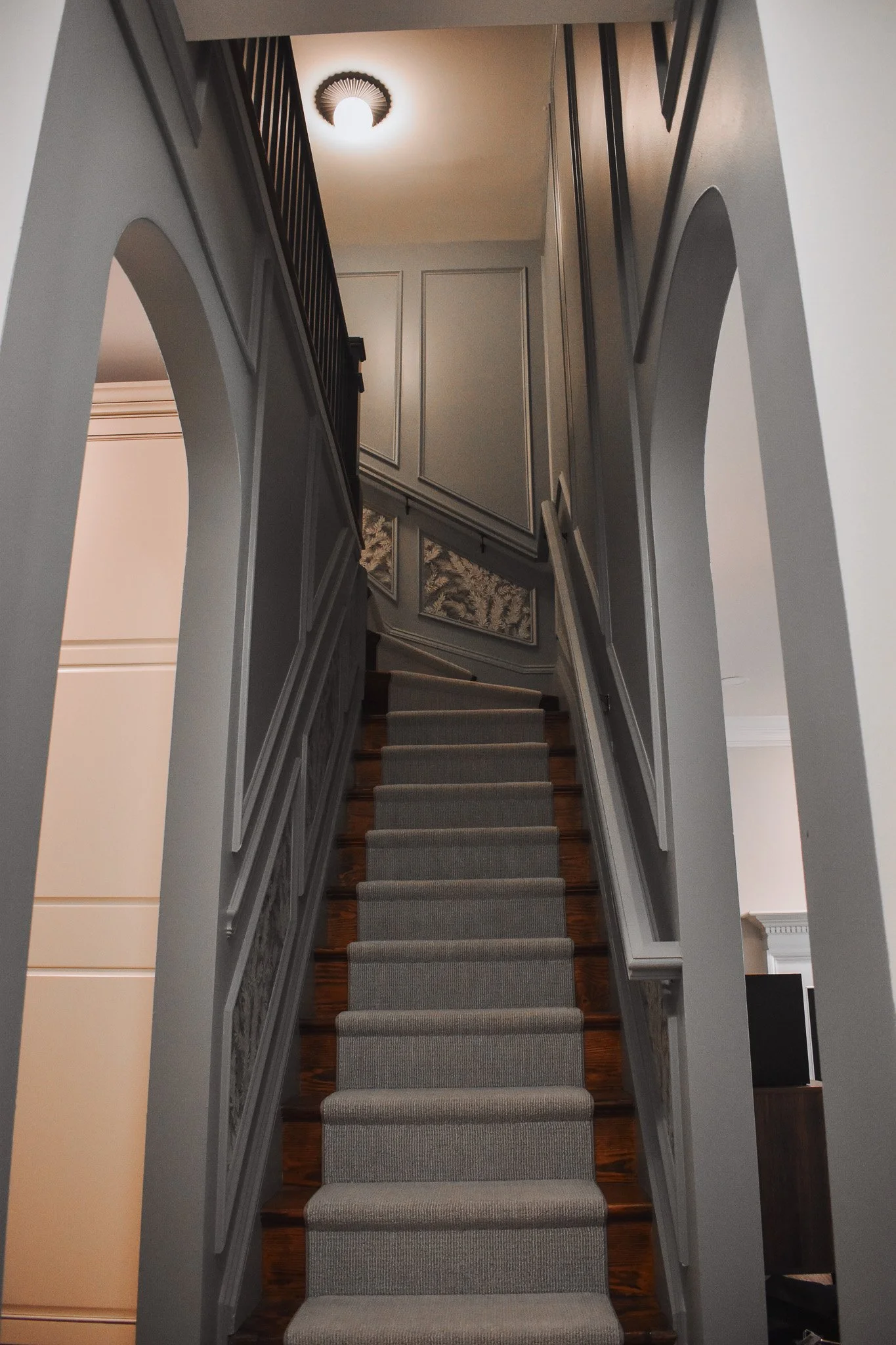 Interior view of a staircase with beige carpeting, dark wooden steps, wood molding, and blue walls, leading to an upper floor with a ceiling light fixture, wallpaper, picture frame molding