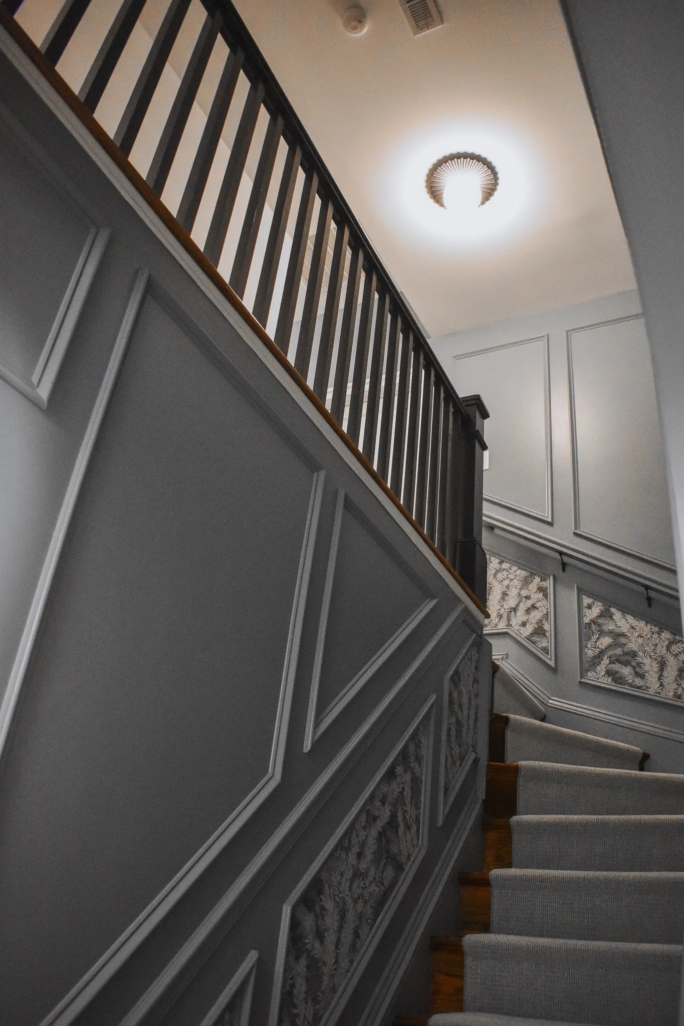 Photo of a staircase with gray and white decorative wall paneling, a wooden handrail, and a ceiling light fixture.