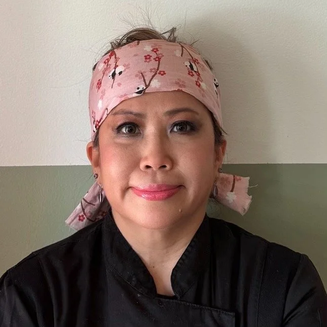 Woman in a black chef coat and pink bandana with cherry blossom pattern, standing against a plain wall.