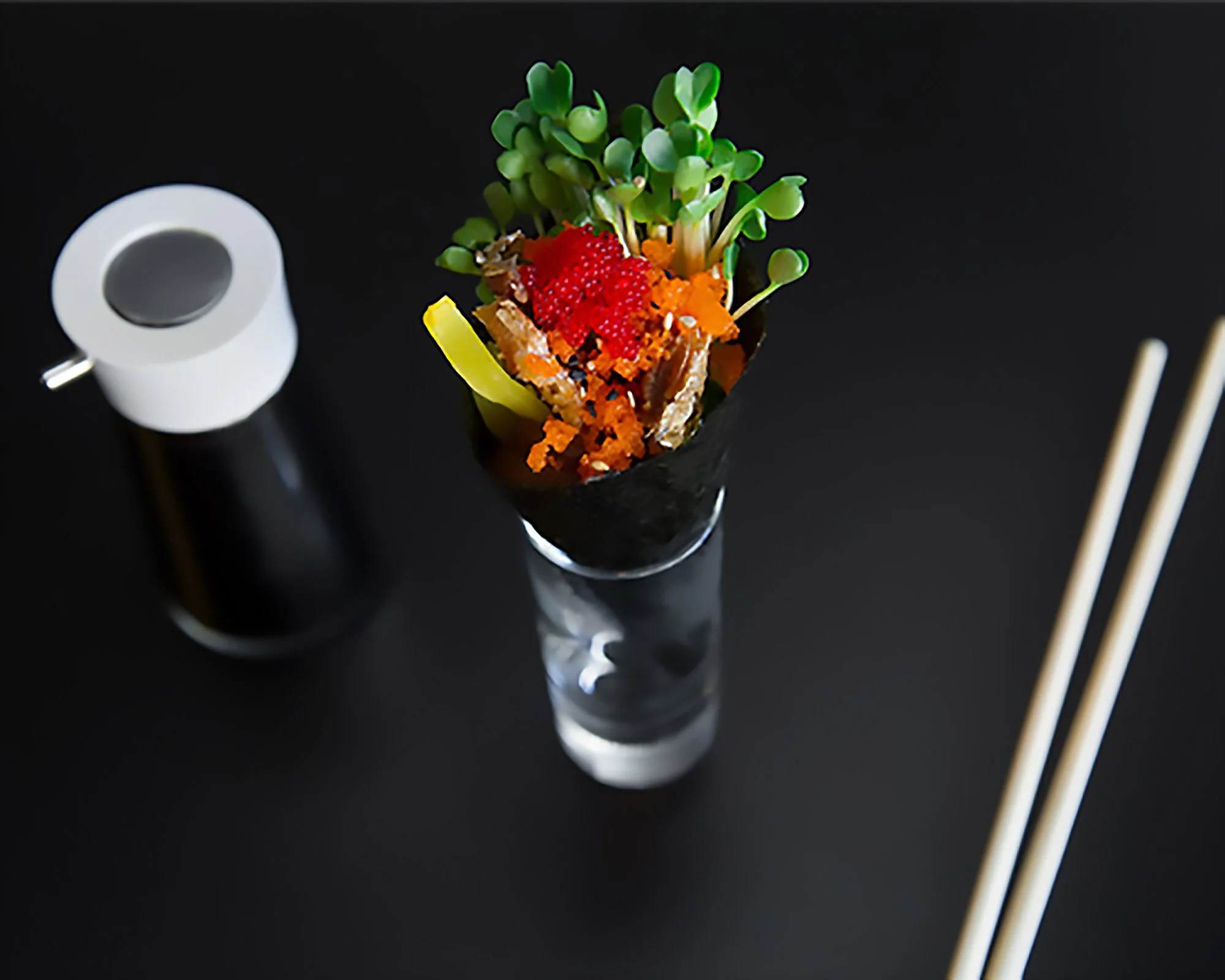 A serving of sushi with microgreens, fish, and roe in a glass, chopsticks, and soy sauce on a black surface.