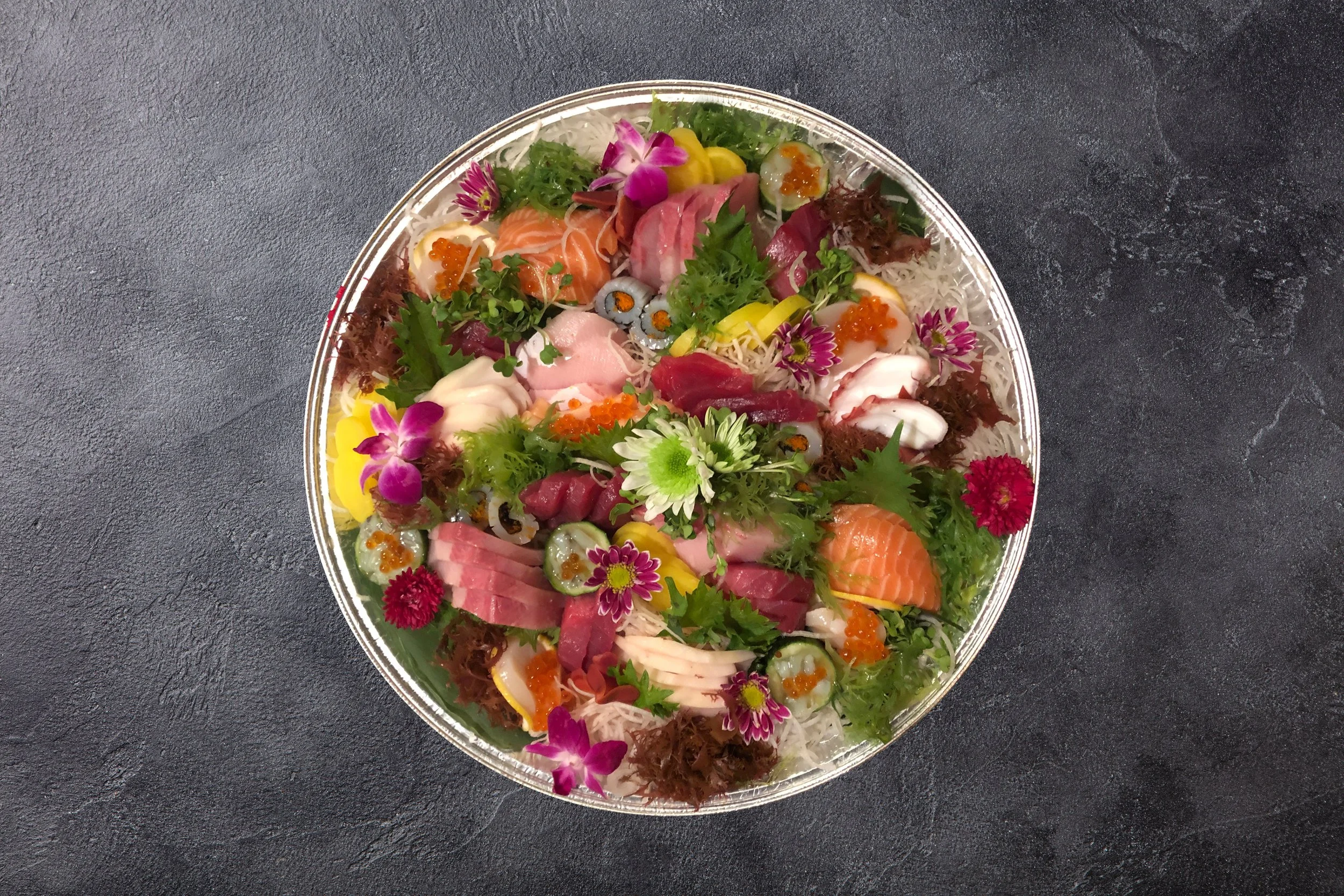 A round platter of assorted sashimi and garnishes, including slices of raw fish, salmon, tuna, and yellowtail, topped with edible flowers, fish roe, and green herbs, set on a dark textured surface.