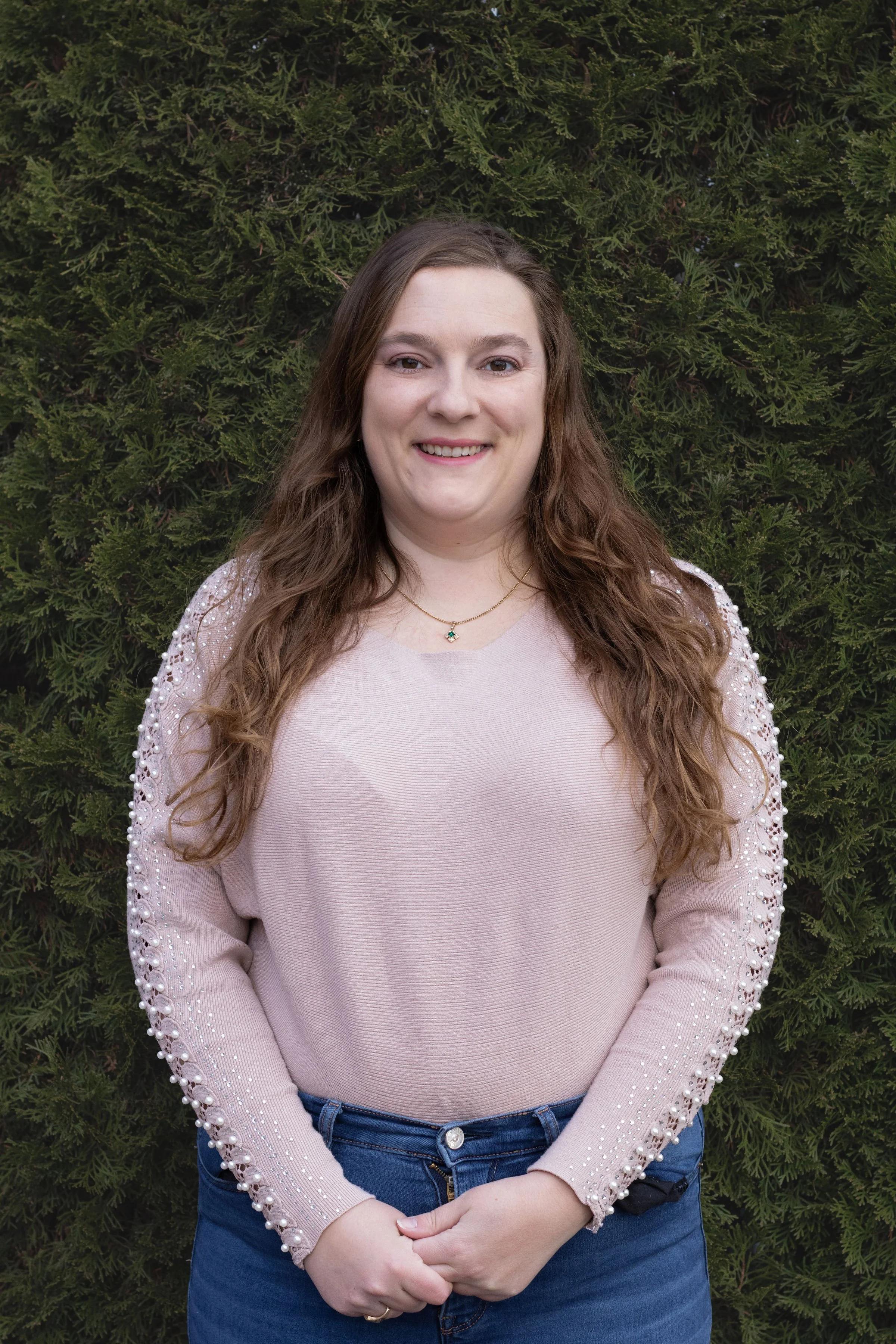 A woman with long wavy brown hair smiling, standing outside in front of green foliage, wearing a light pink sweater with pearl embellishments on the sleeves and blue jeans.