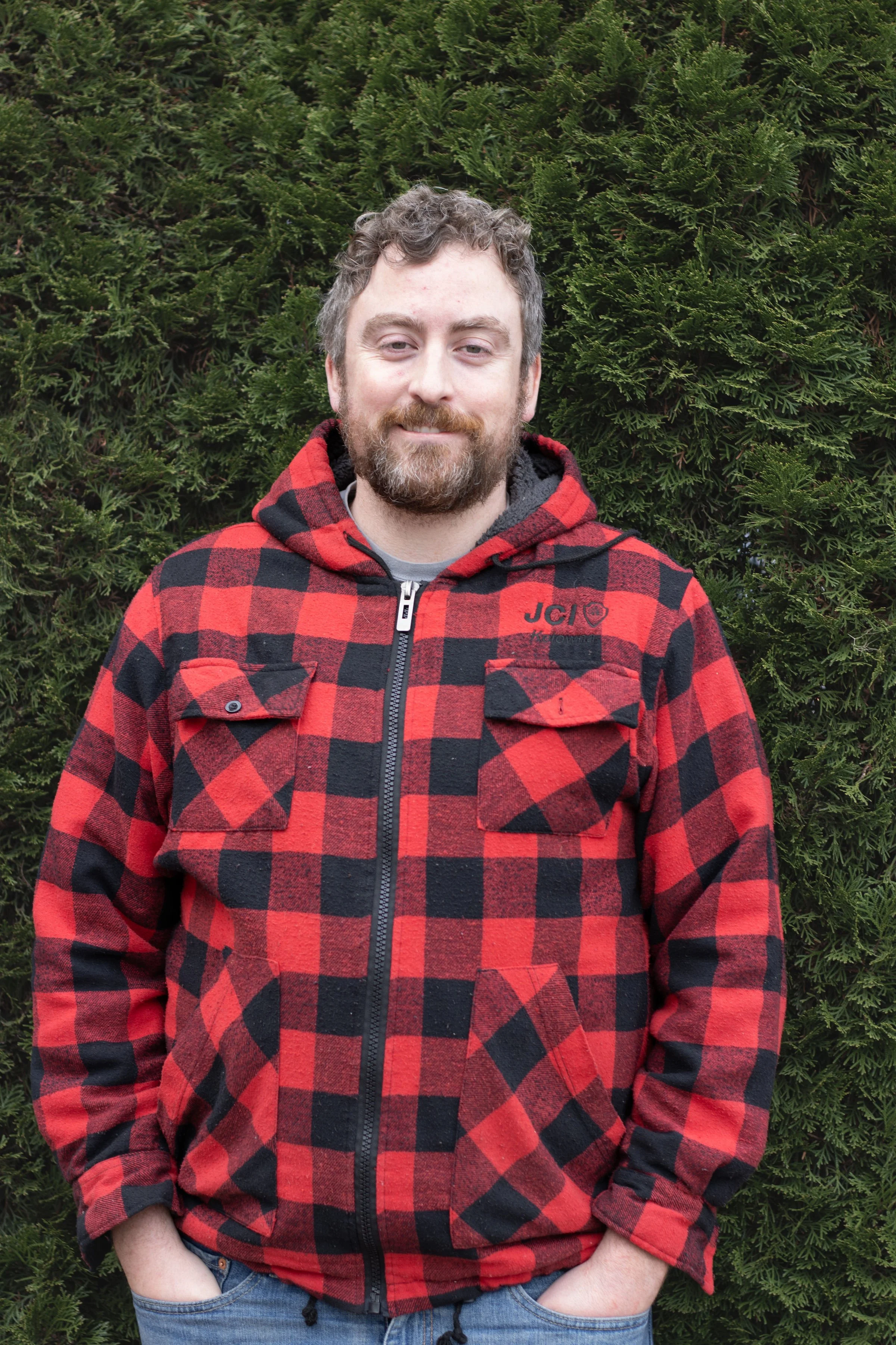 A man with a beard and curly hair standing outdoors in front of green bushes, wearing a red and black checkered jacket with a hood.