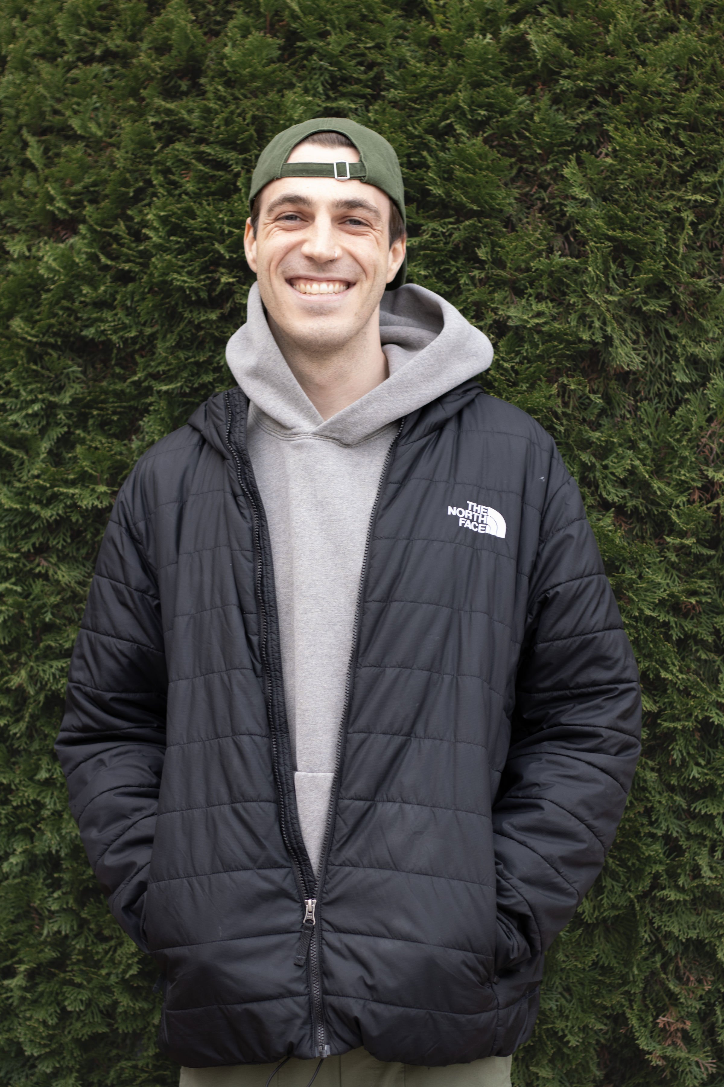 A young man wearing a black The North Face puffer jacket, gray hoodie, and green cap, smiling and standing in front of green pine trees.