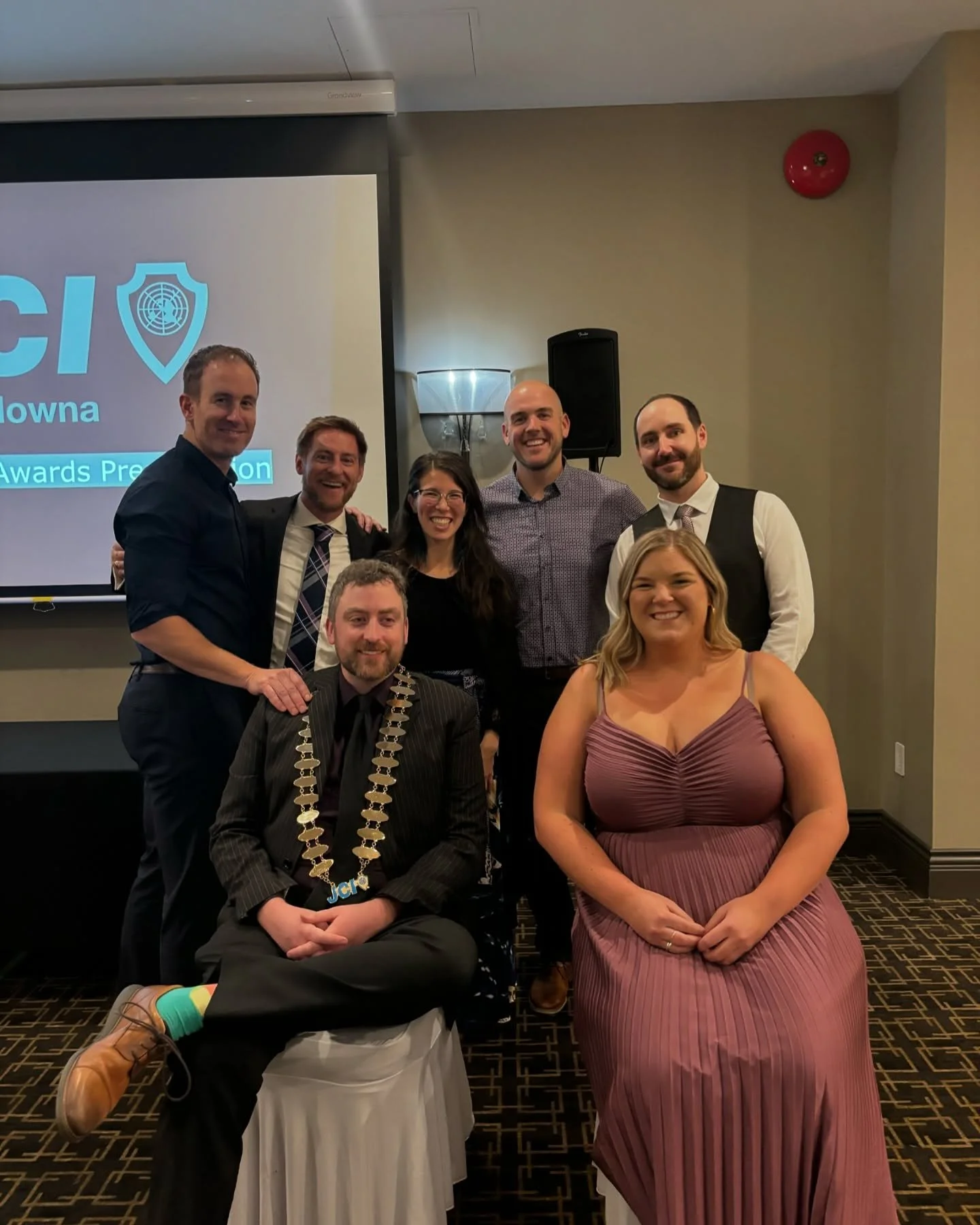 Group of people at an award ceremony, with some sitting and others standing, in a hotel conference room.