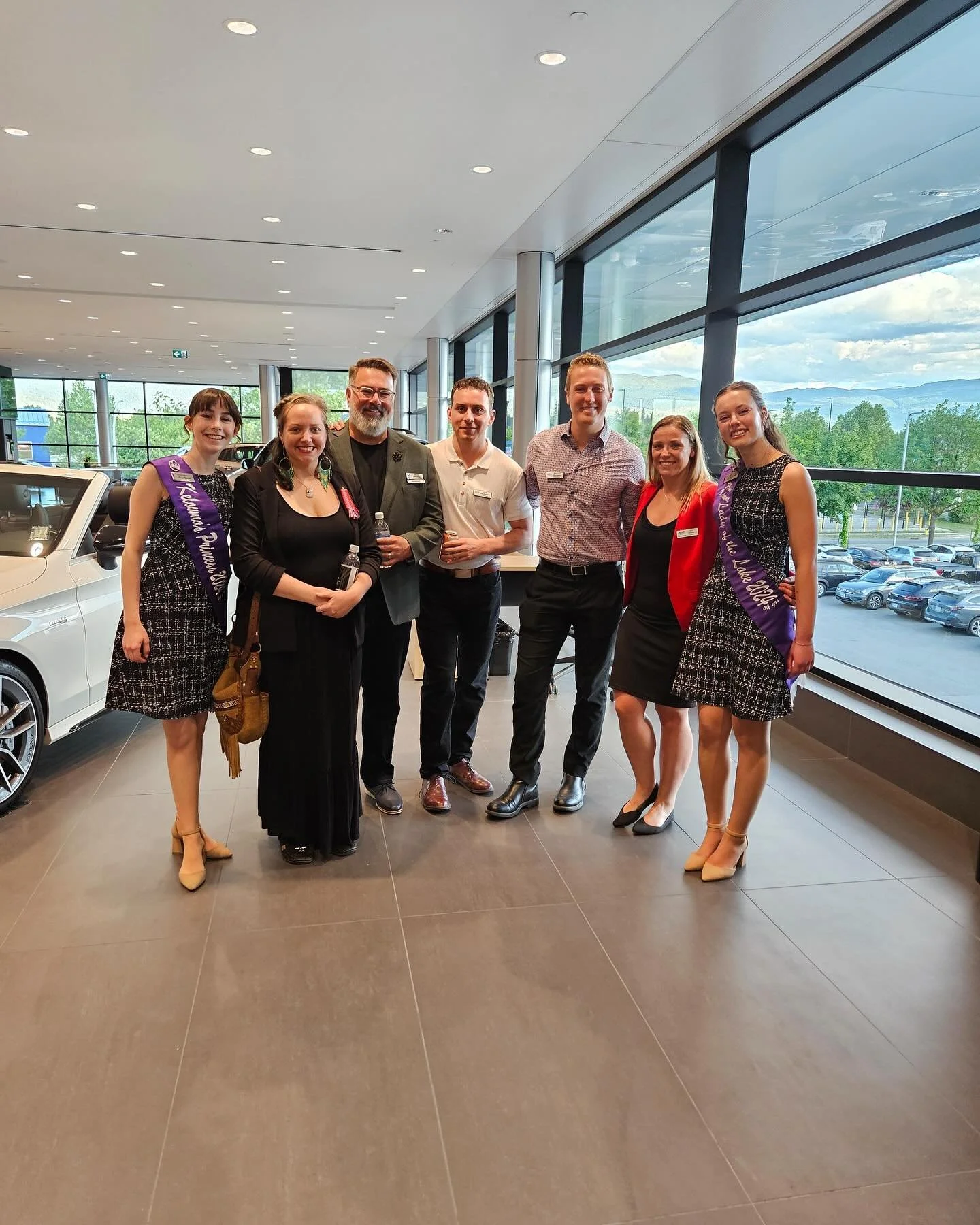 Group of seven people standing inside a car dealership showroom with large windows showing cars and a parking lot outside.