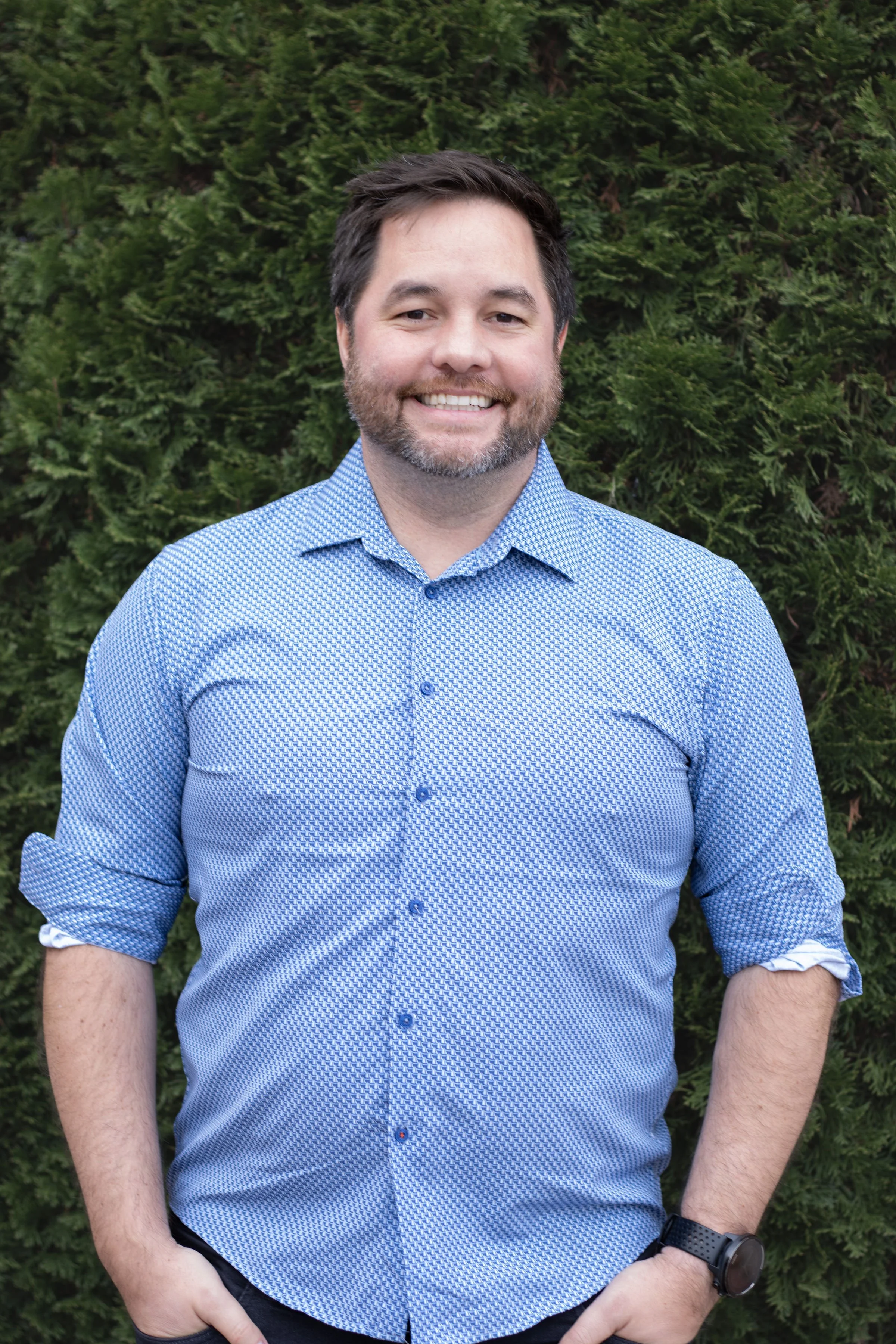 A man smiling outdoors in front of green foliage, wearing a blue patterned shirt with the sleeves rolled up and a black watch.