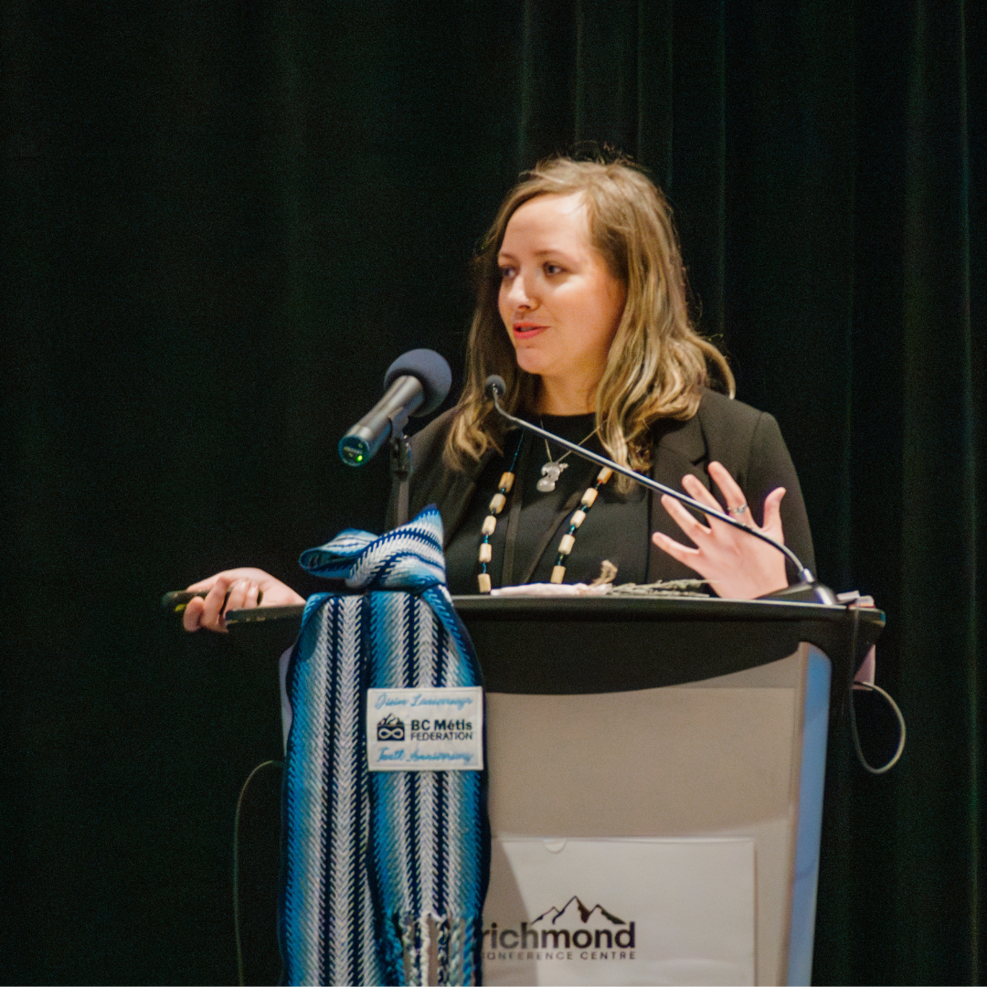 A woman giving a presentation at a podium with a BC Metis Federation banner and a blue woven scarf hanging on the side.
