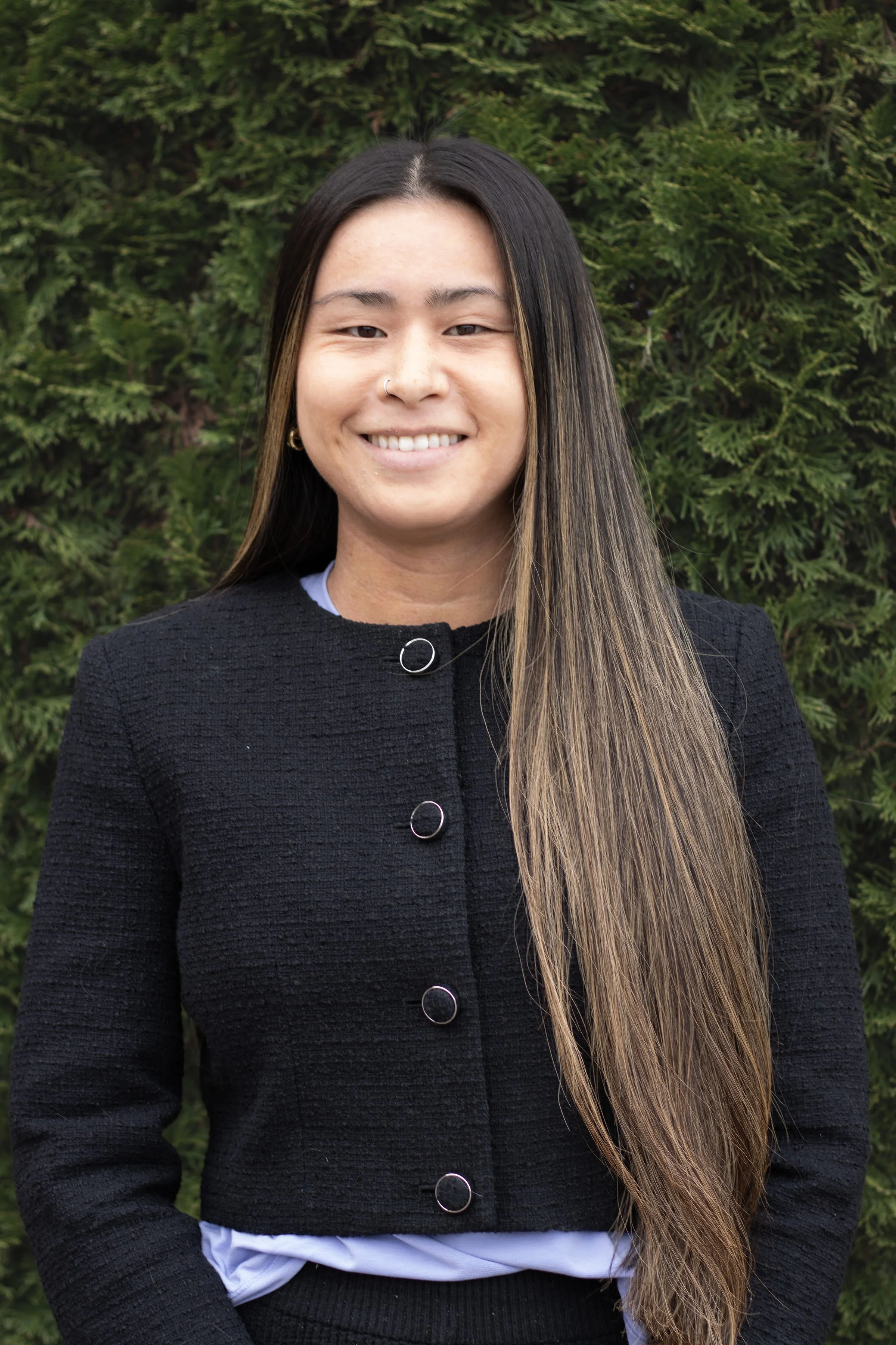 A young woman with long straight hair, wearing a black textured jacket with metal buttons, standing outdoors in front of green bushes, smiling at the camera.