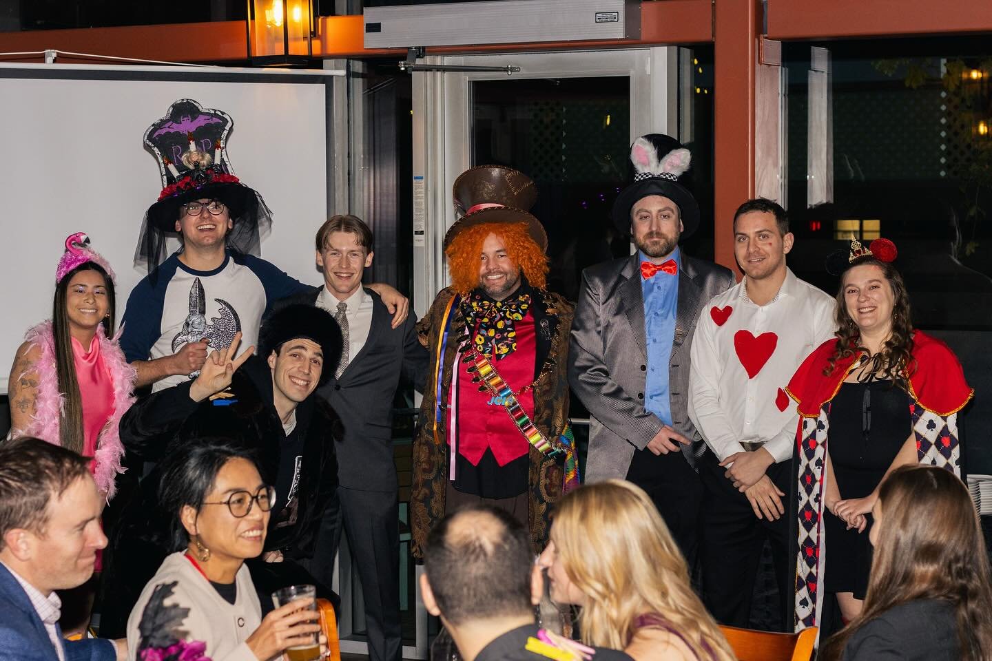 Group of people dressed in colorful costumes, celebrating at a party or event, with some wearing hats and accessories, in a decorated indoor space.