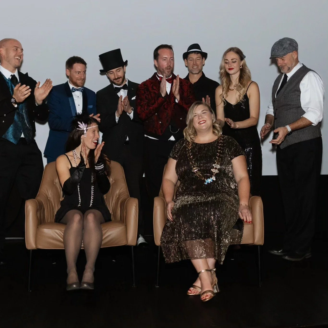 Group of people dressed in vintage clothing, some sitting on chairs and others standing behind, smiling and clapping, during a celebration or event.