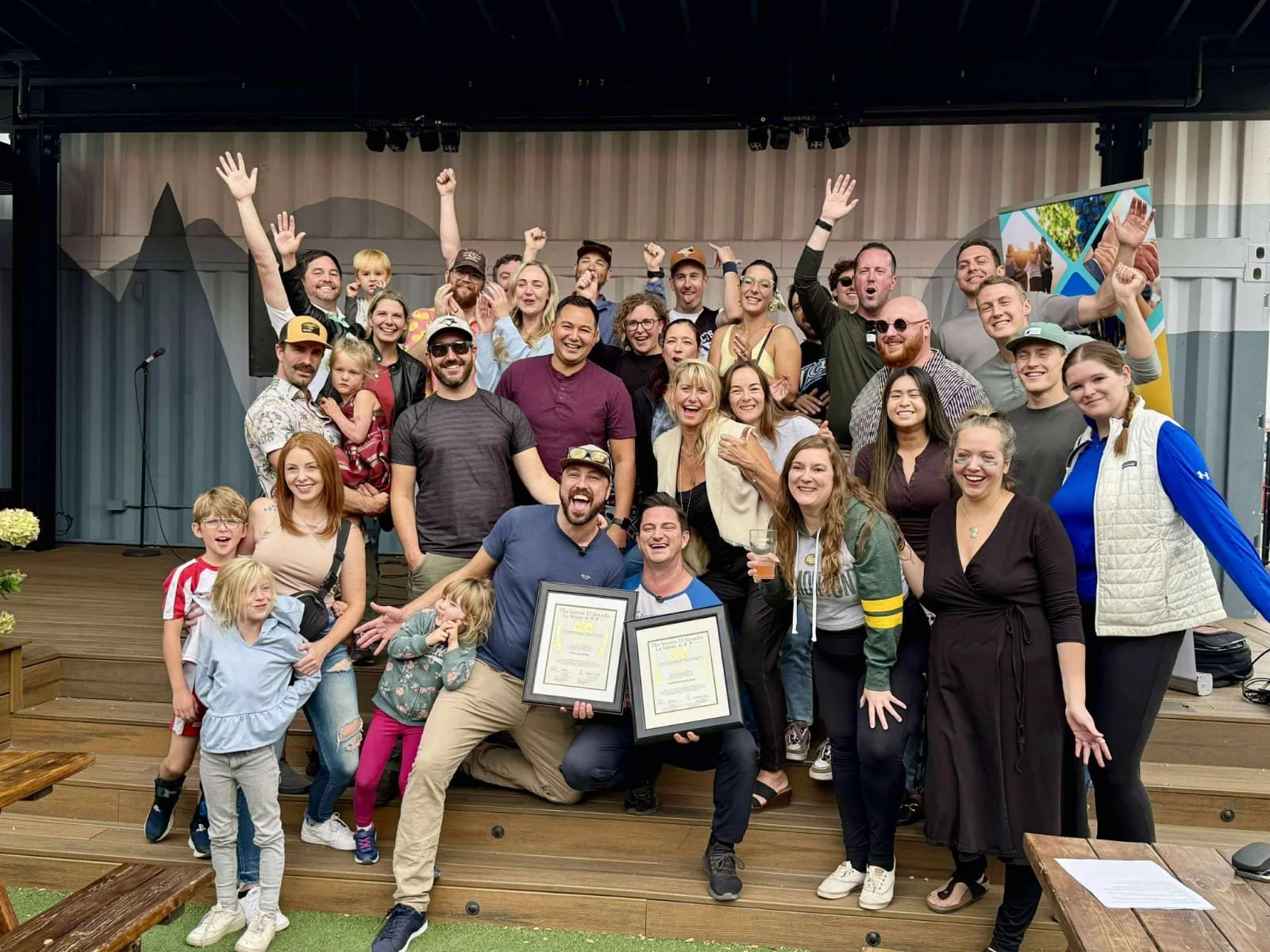 A large, diverse group of people gathered on a wooden stage, celebrating and posing for a photo. Some are holding framed certificates, and many are smiling, cheering, and raising their hands.