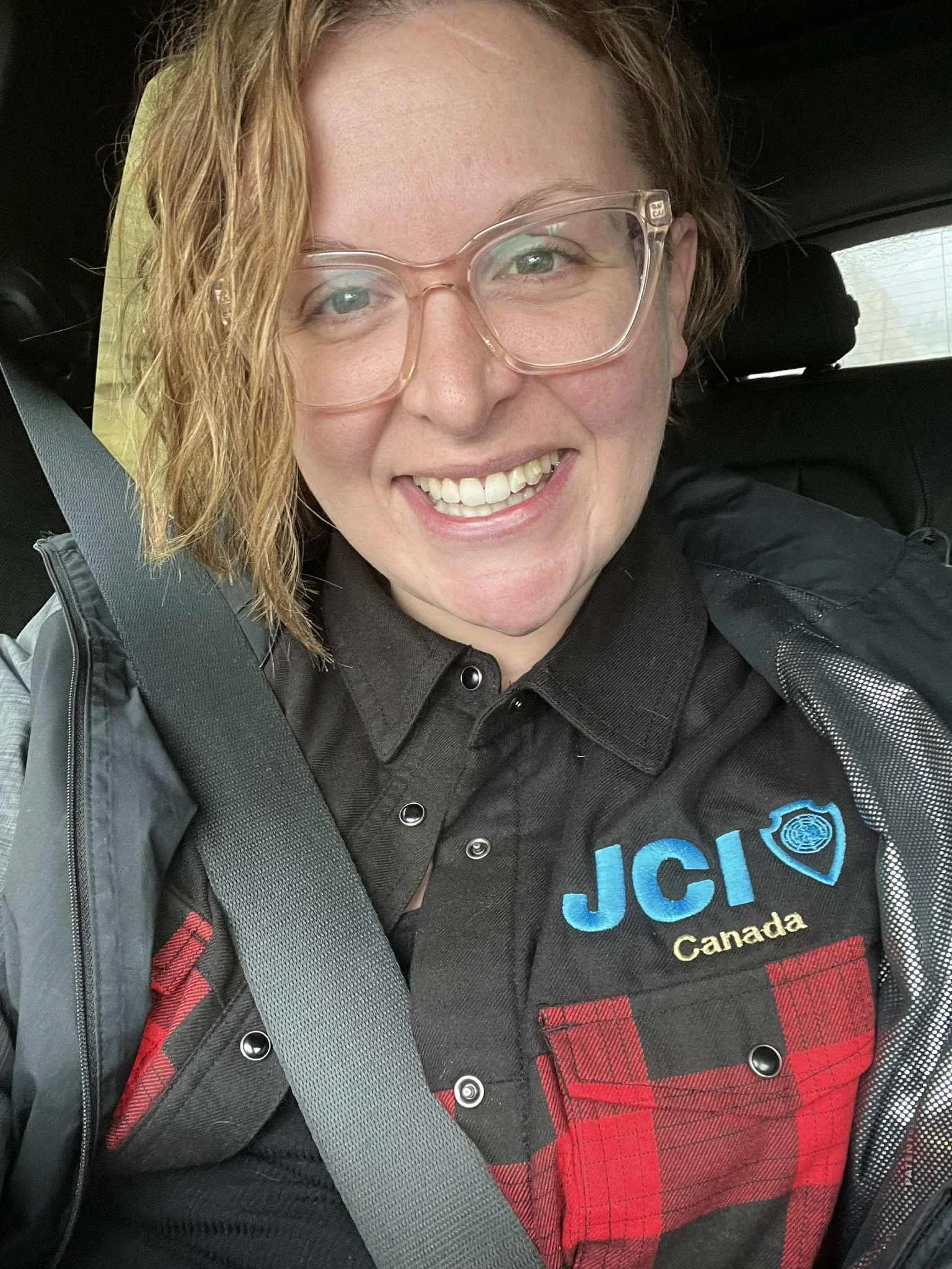 A woman smiling while sitting in a vehicle, wearing glasses and a black and red uniform with JCI Canada logo.